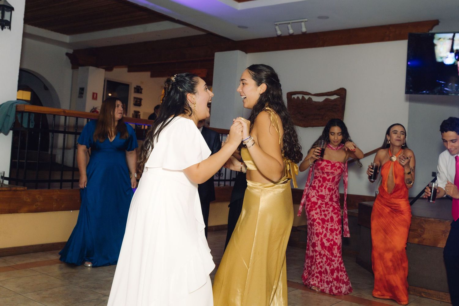 Bride in white dress dances and laughs with a guest in gold dress at reception.