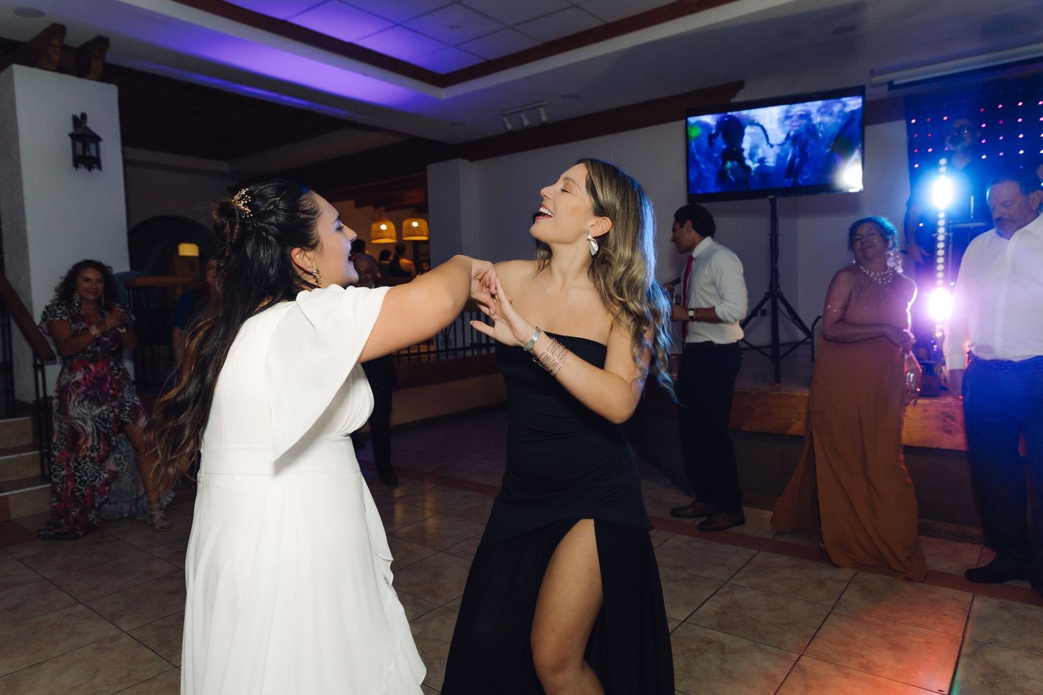 Two women dancing together at a wedding reception, one in white bridal gown and one in black dress.