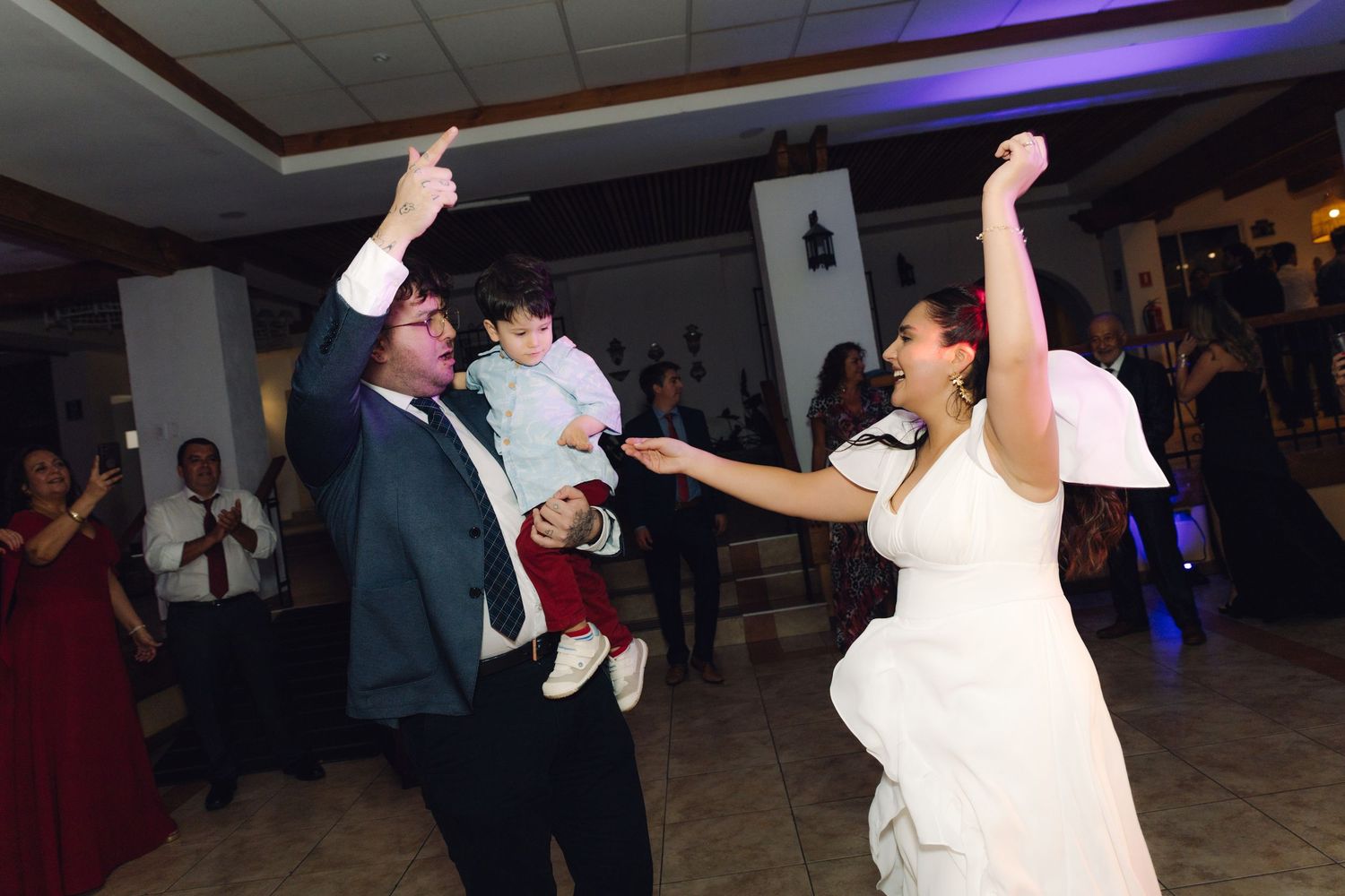 Bride and groom dancing joyfully at wedding reception while groom holds a young child.