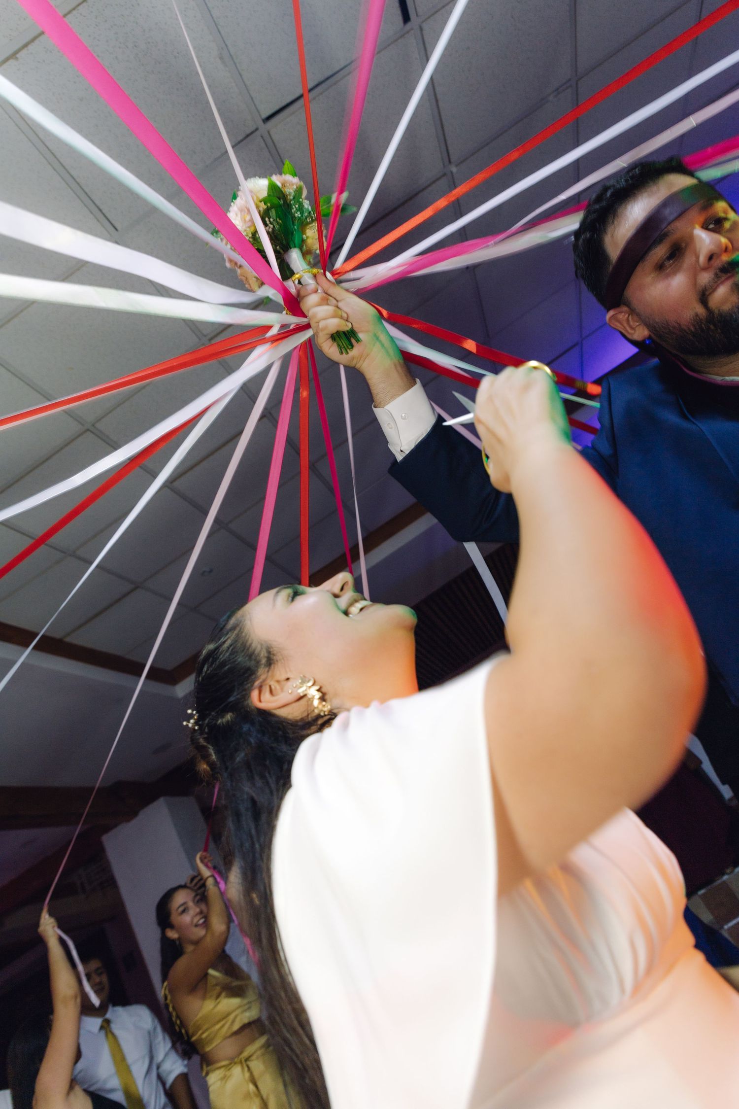 Bride and guest pulling colorful ribbons from wedding bouquet at festive reception celebration.