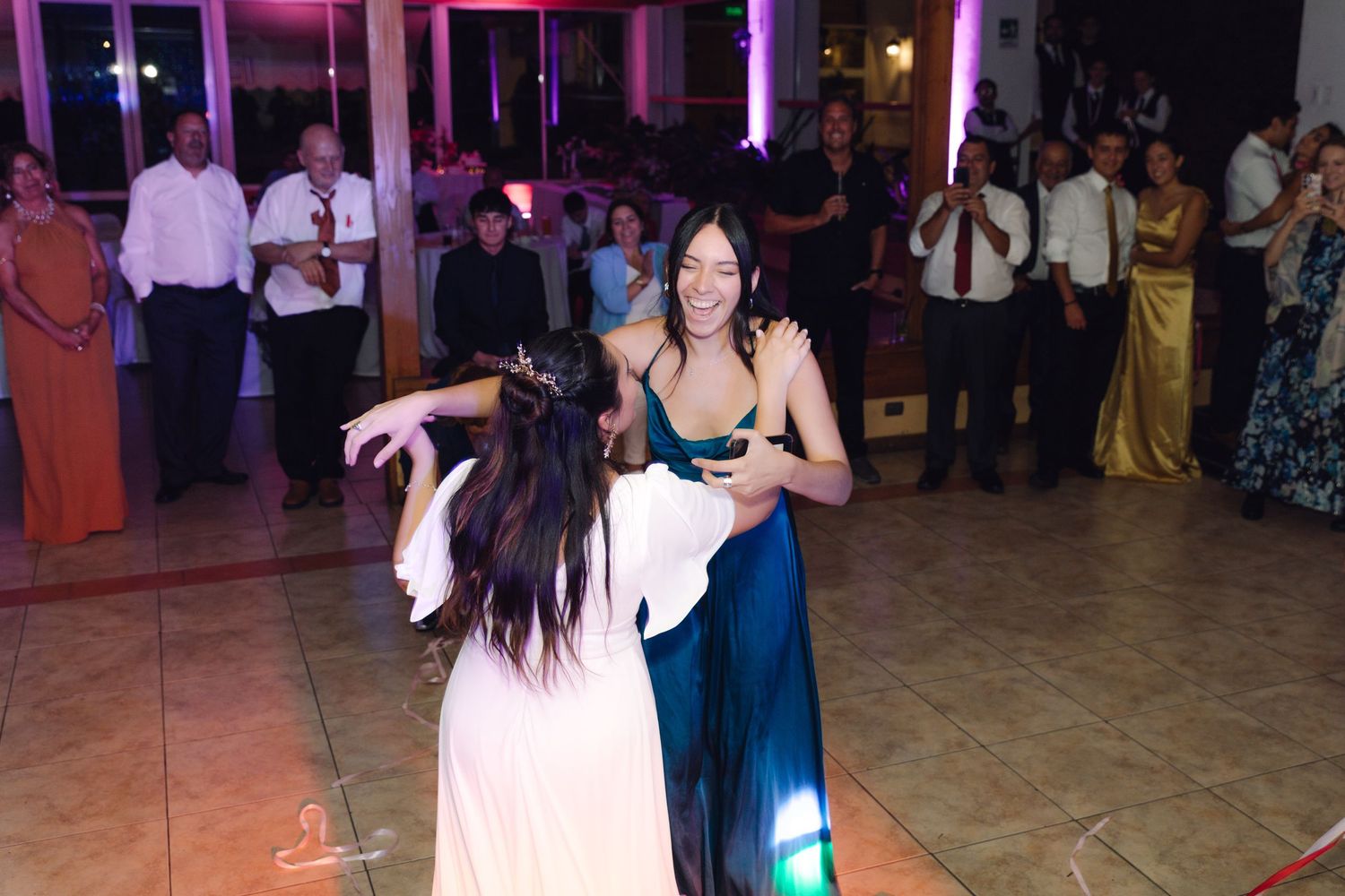 Two women dancing together on wedding reception dance floor with guests watching in background.