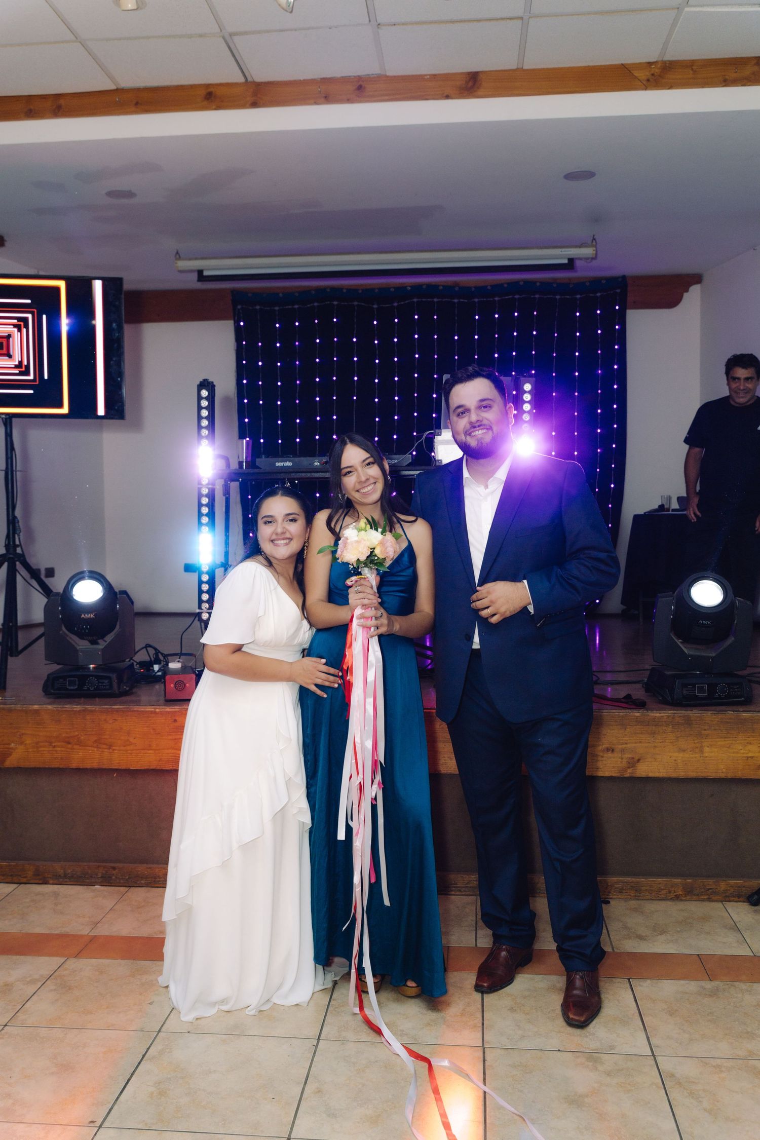 Bride and groom posing with bouquet winner in teal dress in front of DJ stage at wedding.