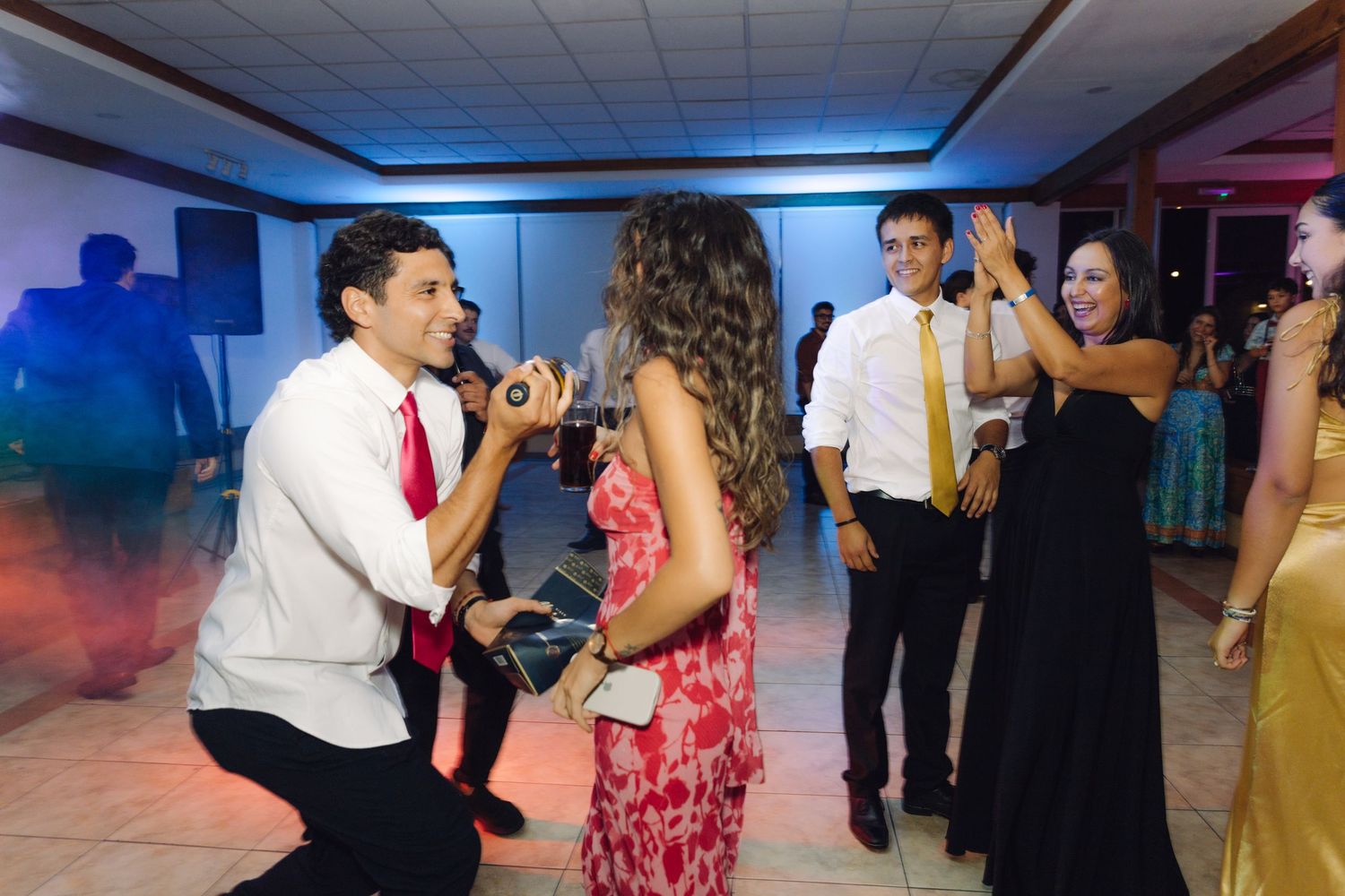 Man kneeling on dance floor serenading woman with microphone at lively wedding reception.
