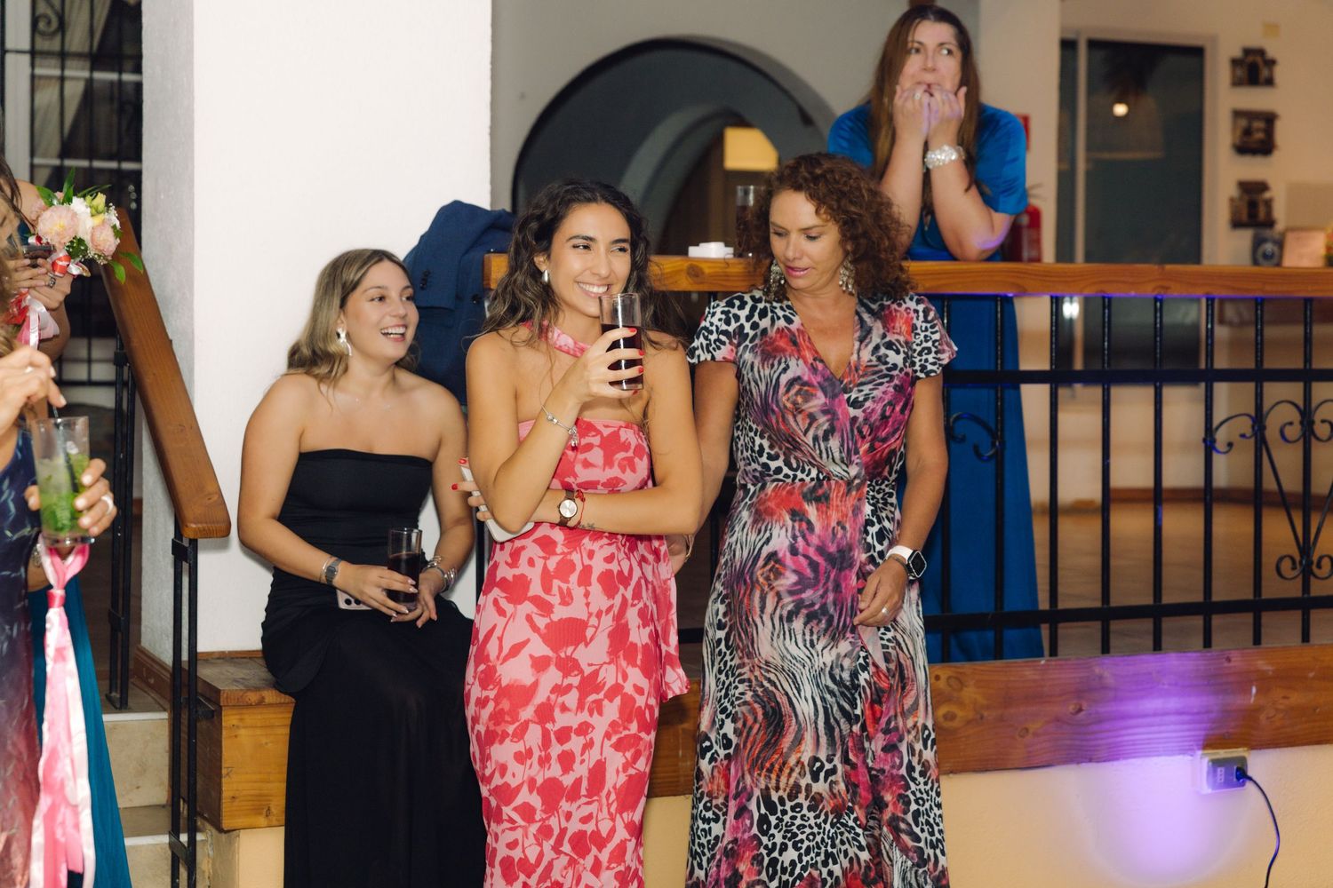 Three women smiling and holding drinks at wedding reception near decorated staircase railing.