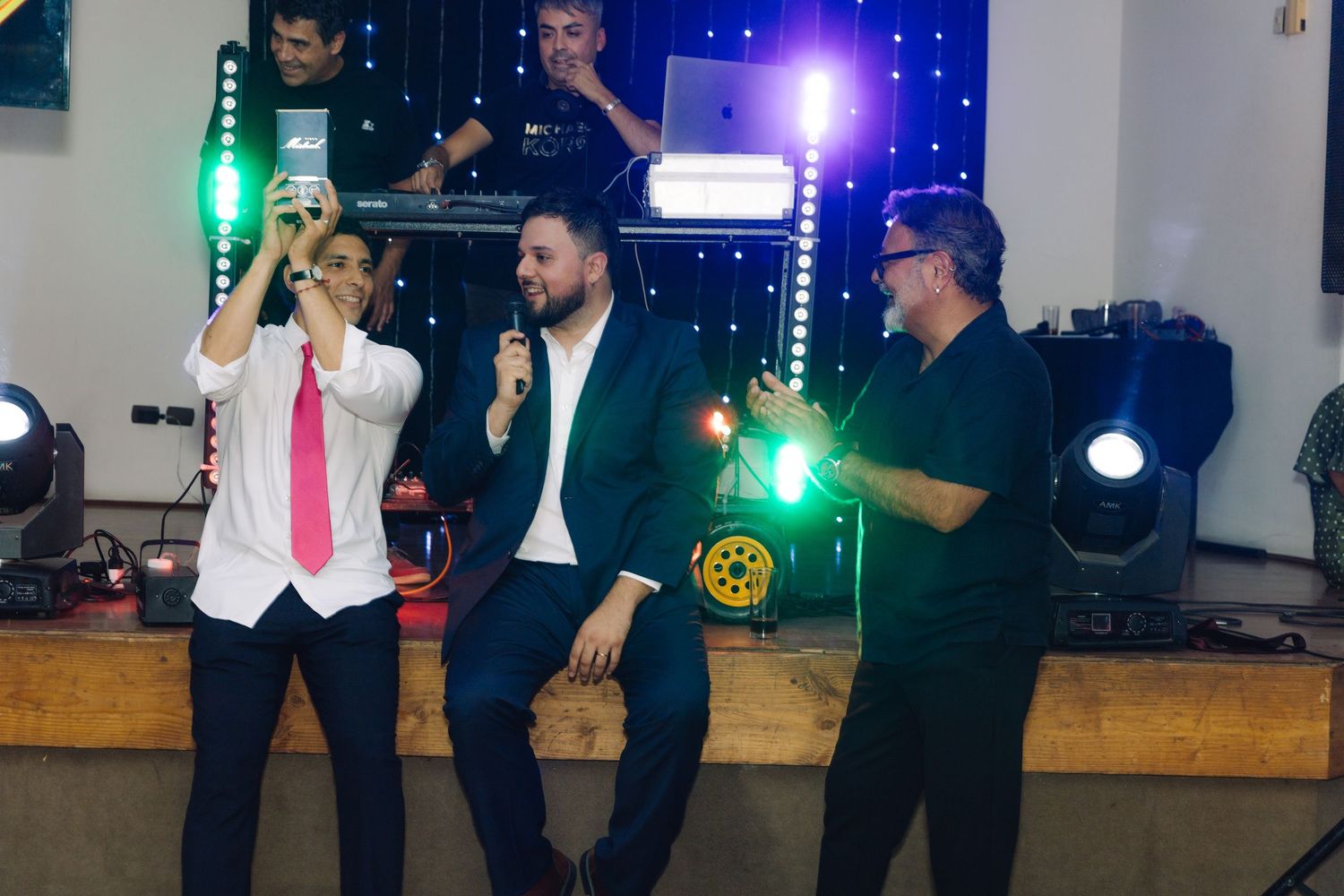 Three men performing on stage with microphones at a wedding reception with DJ equipment.