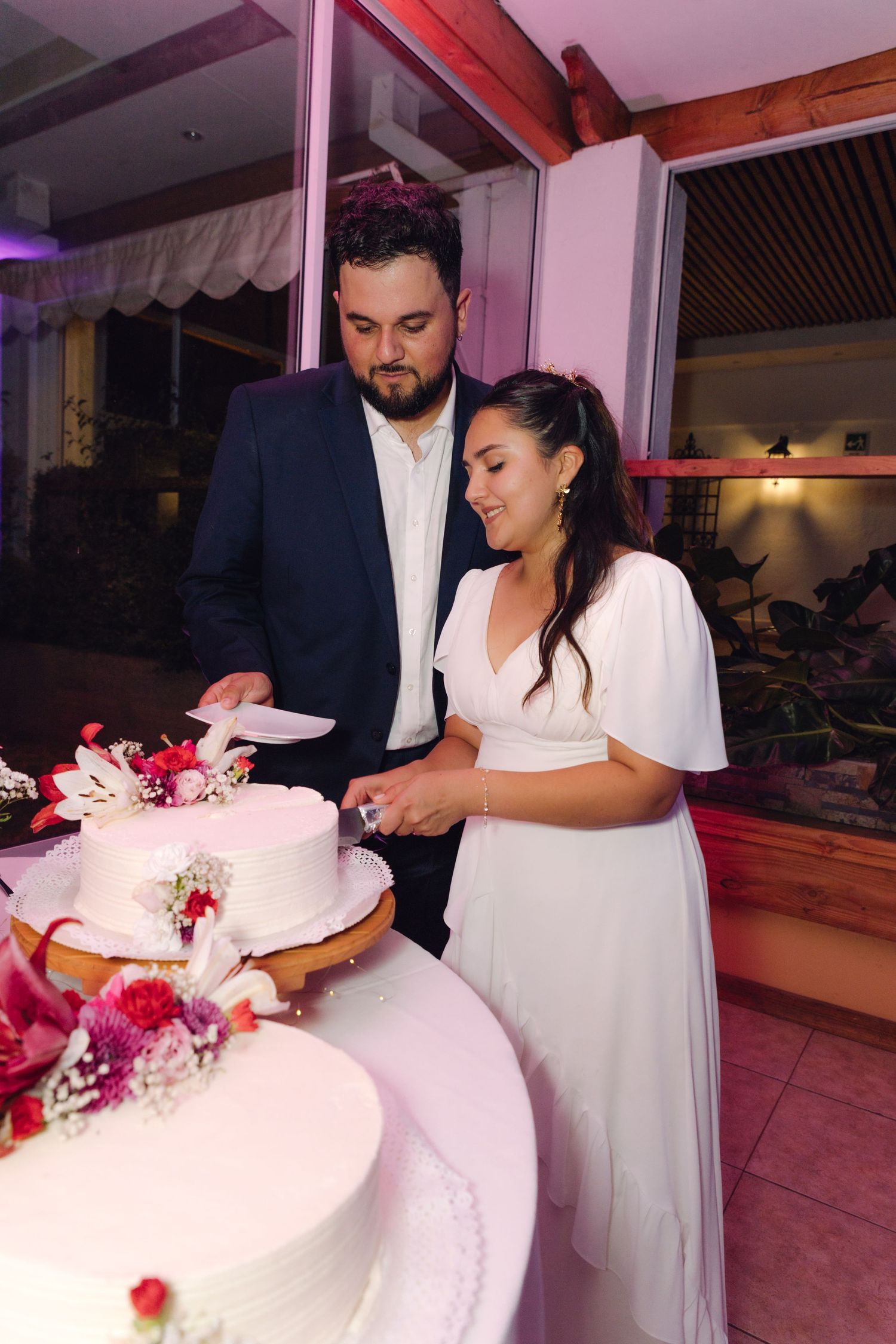 Smiling couple in wedding attire cutting a white tiered cake at their reception.