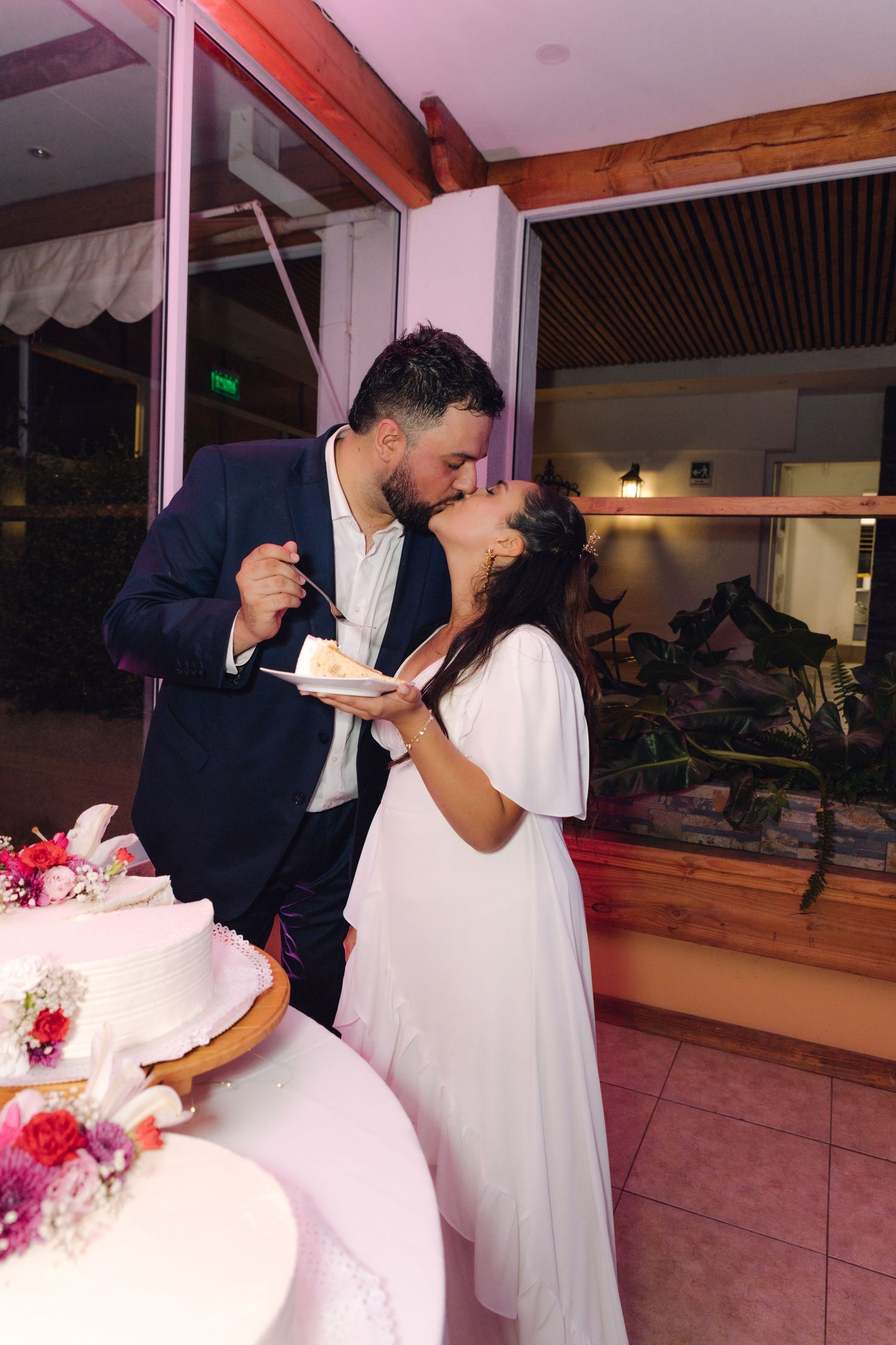 Newlyweds sharing a romantic kiss while holding a plate of wedding cake.