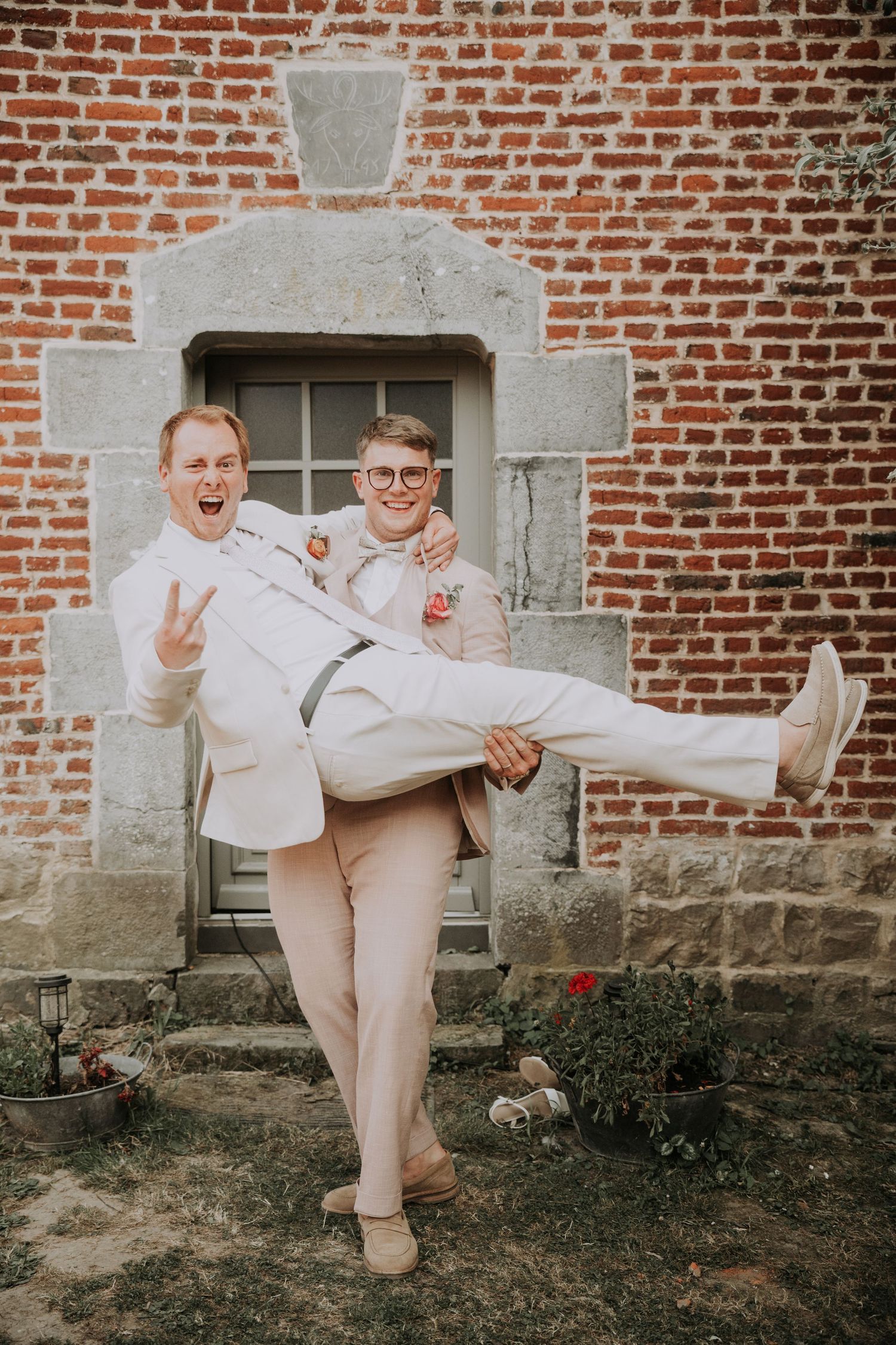Two grooms celebrate joyfully, one carrying the other outside a rustic brick building on their wedding day.