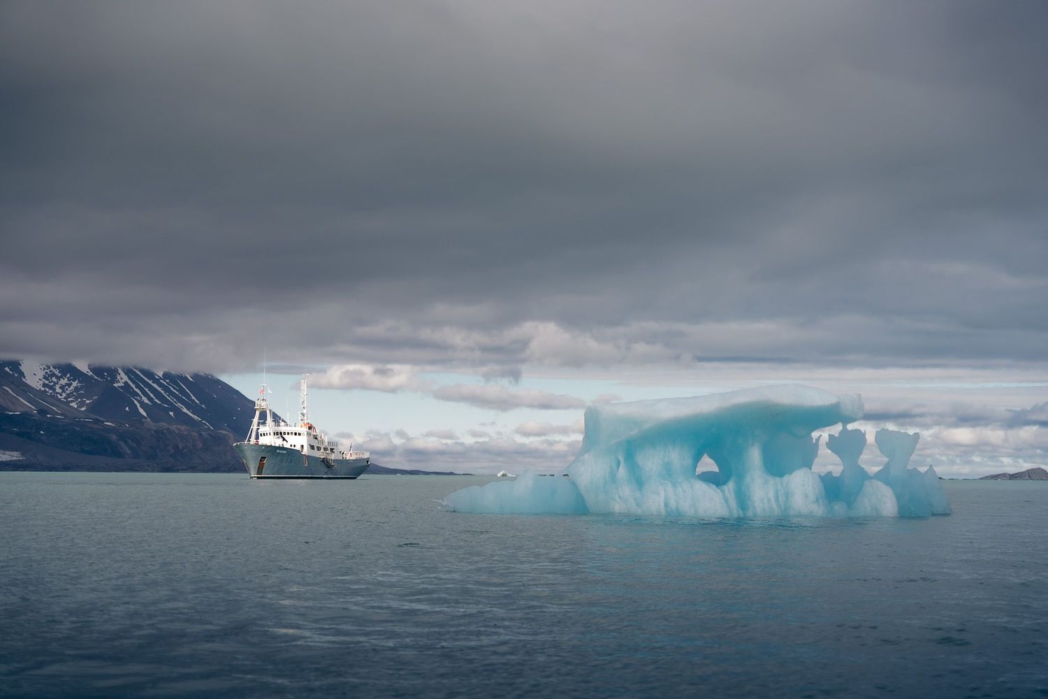Le Polarfront et un iceberg percé d'arches sous une lumière d'orage plombée