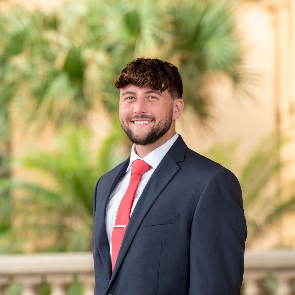 Young professional man in navy suit and red tie smiling outdoors with palm trees in background. Professional Headshot.