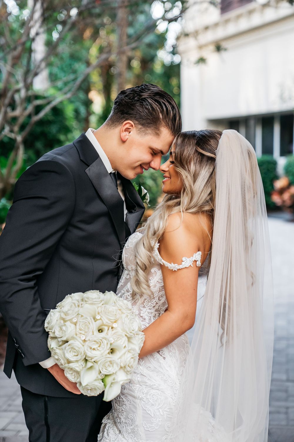 Bride and groom touching foreheads tenderly outdoors, she holds white rose bouquet, wearing lace gown with veil, he in black tux.