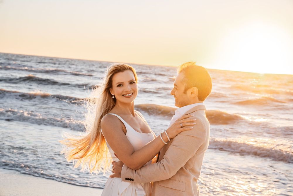 Couple embracing on a beach at golden sunset, woman in white dress smiling, waves in the background.