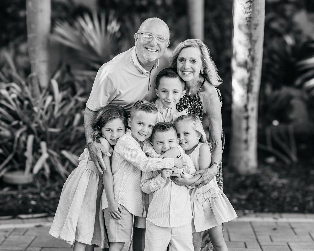 Black and white family portrait of parents with five young children hugging outdoors among tropical palms.