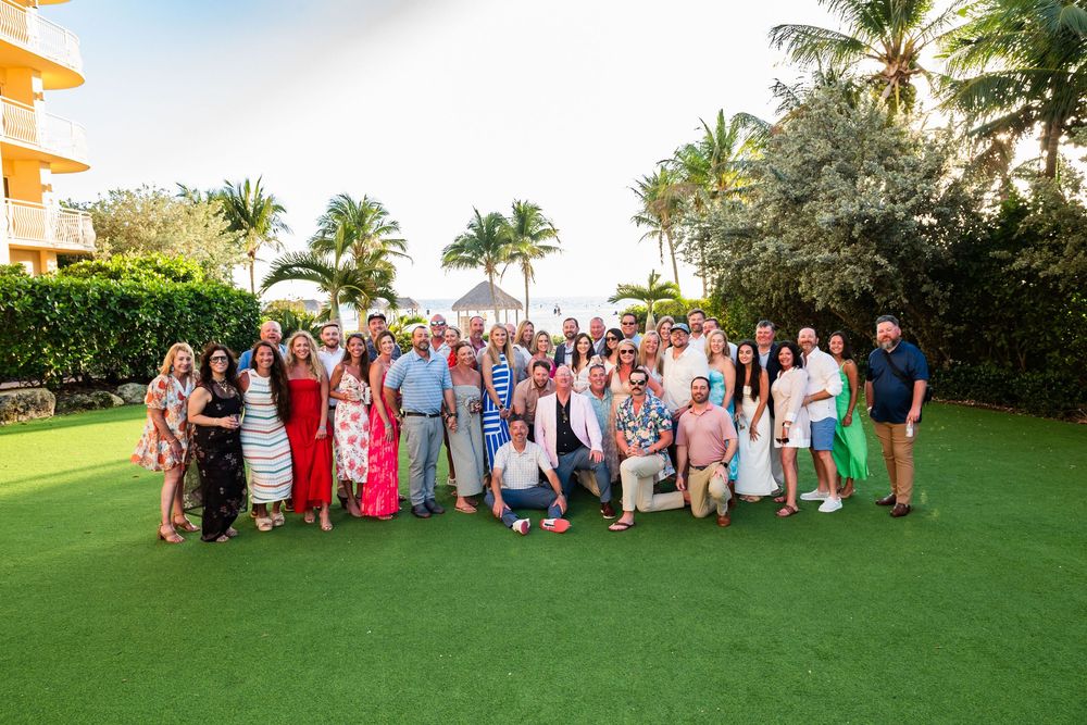Large group of people posing together on a tropical resort lawn with palm trees and ocean views.