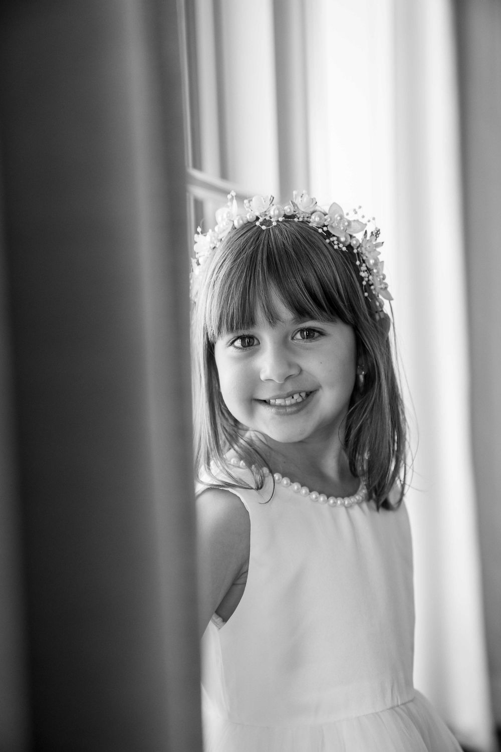 Black and white portrait of a smiling young girl wearing a floral crown and white dress near a window on her aunt's wedding day.