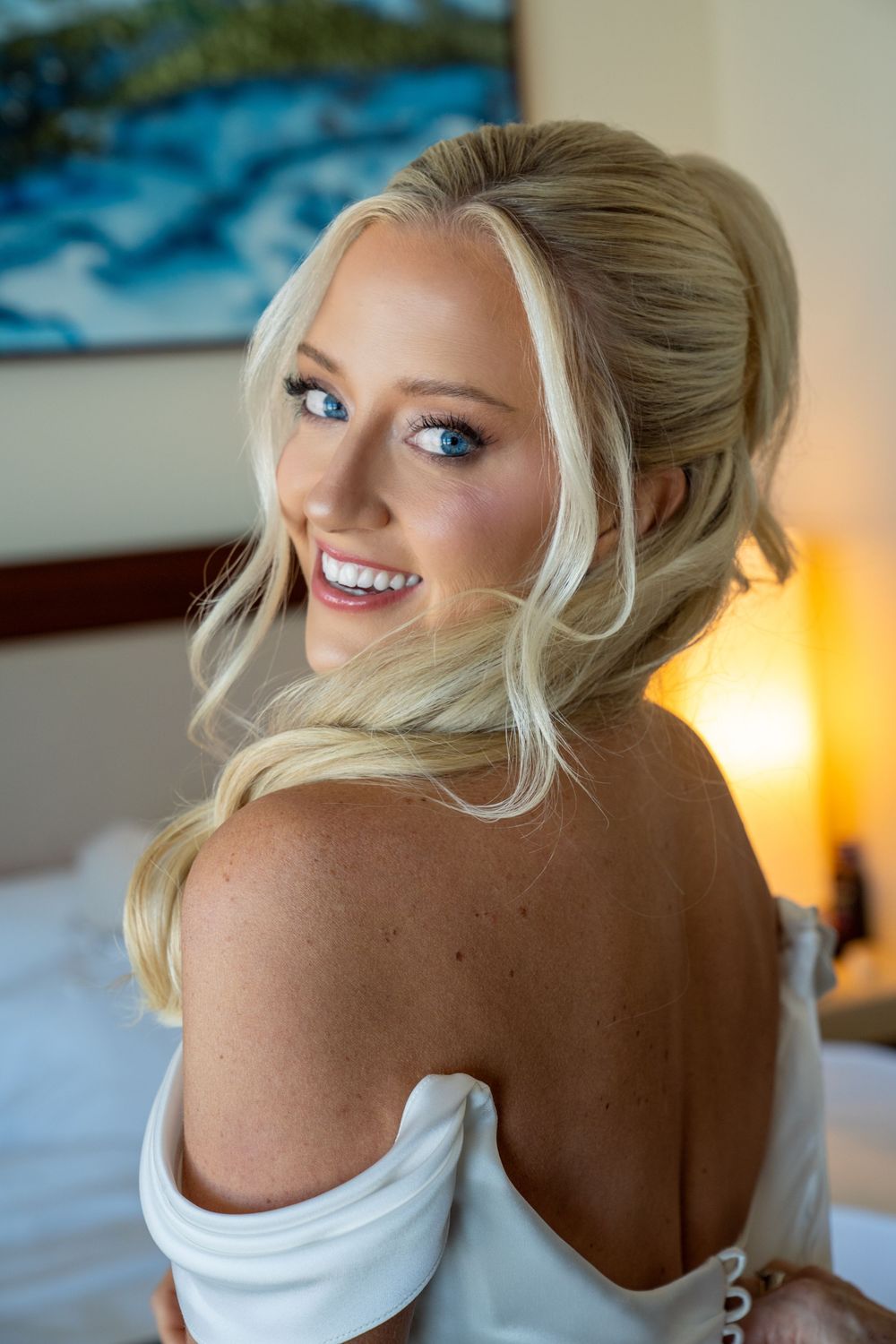 Smiling blonde bride in off-shoulder white gown with elegant updo hairstyle and blue eyes, posing in a warmly lit room.