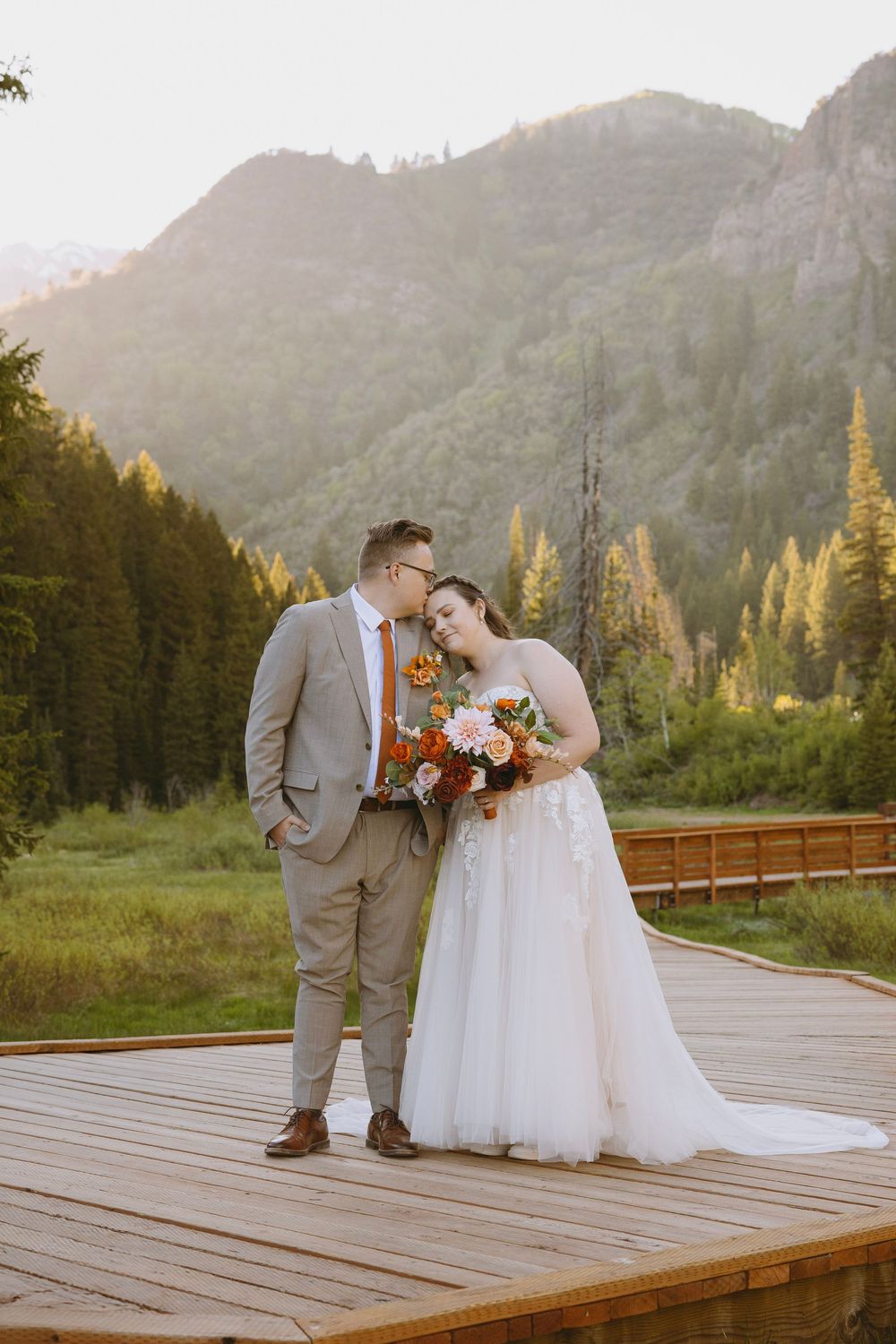 Bride and groom stand on wooden deck with mountain forest backdrop, bride holding autumn bouquet in white gown, groom in tan suit.
