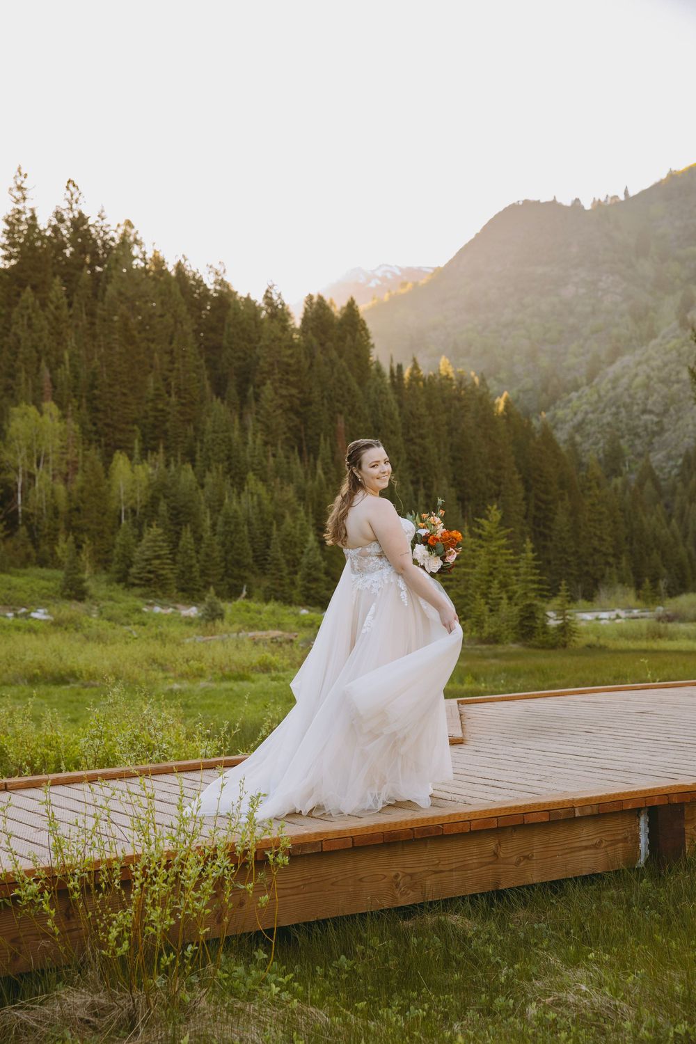 Bride in white gown holding bouquet on wooden deck with mountain forest backdrop at golden hour.
