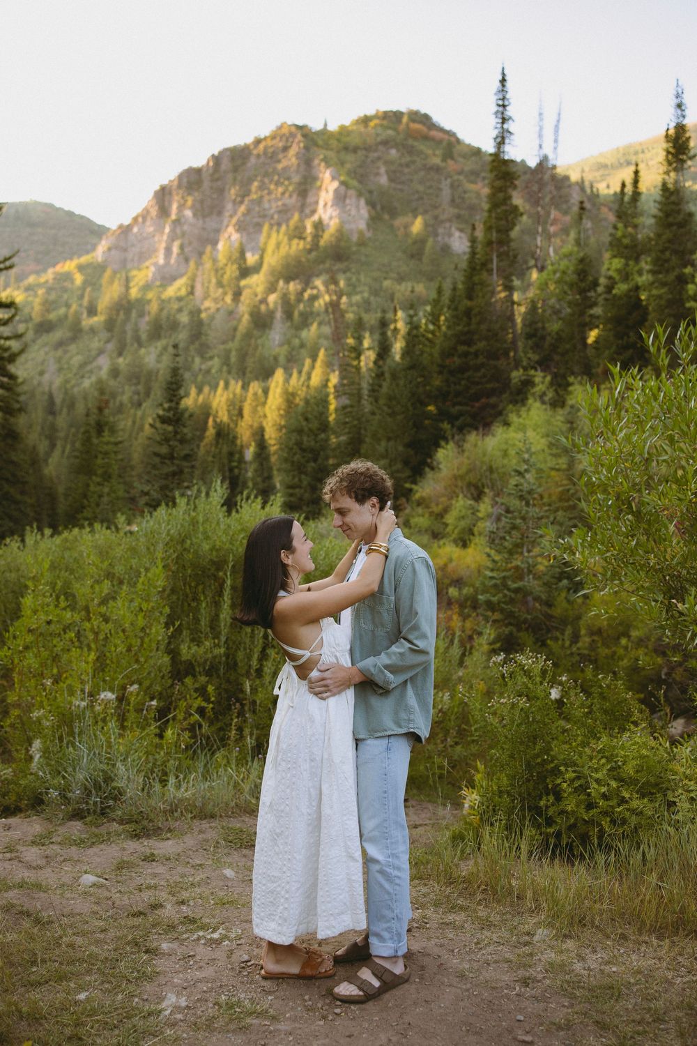 Couple embracing on a mountain trail surrounded by lush green trees and rocky peaks during golden hour.