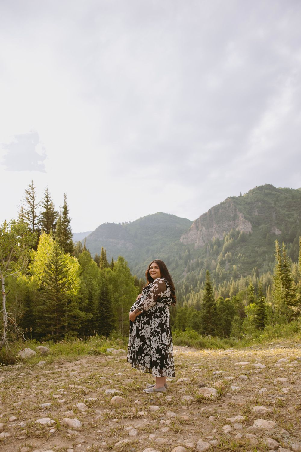 Pregnant woman in floral dress posing on rocky mountain trail surrounded by evergreen trees and scenic peaks.