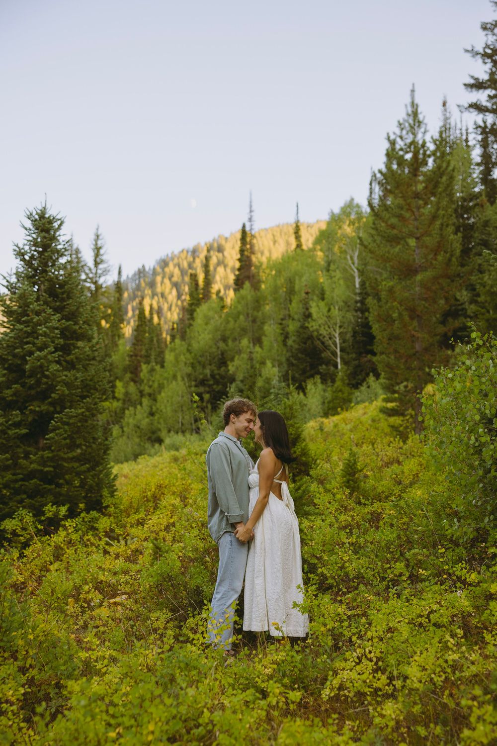 Couple embracing in lush mountain forest clearing, woman in white dress, man in gray suit, golden hillside backdrop.