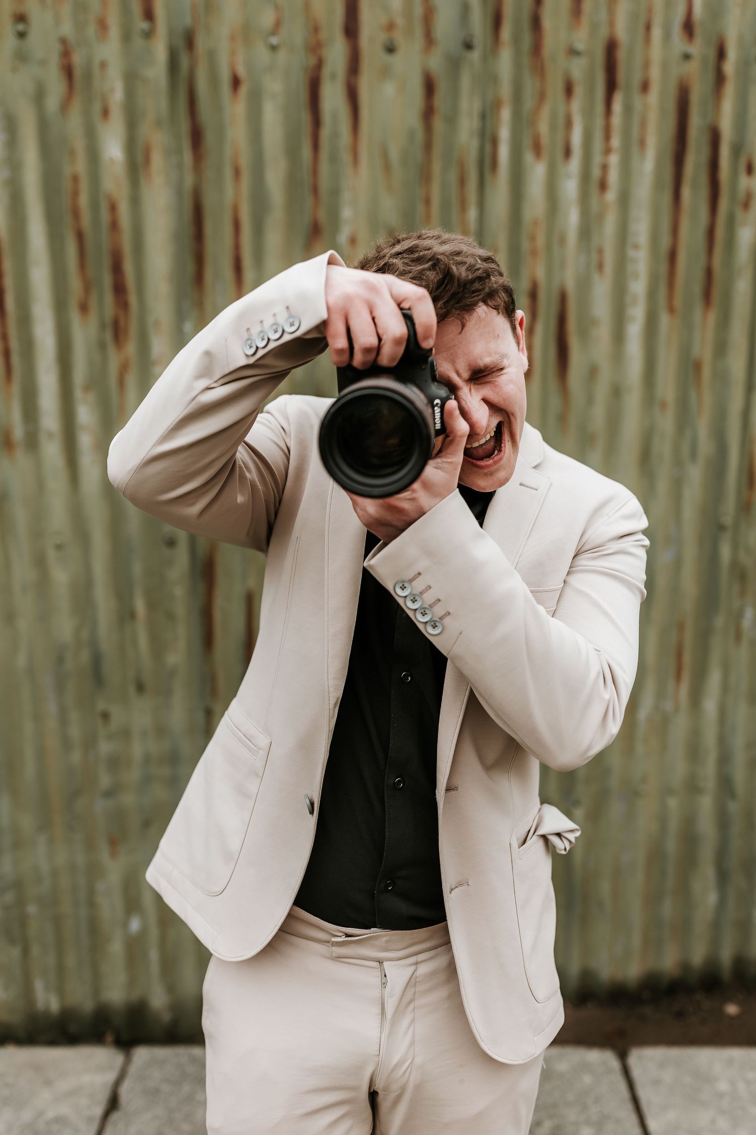 Laughing photographer in cream suit captures photos with professional camera outdoors.