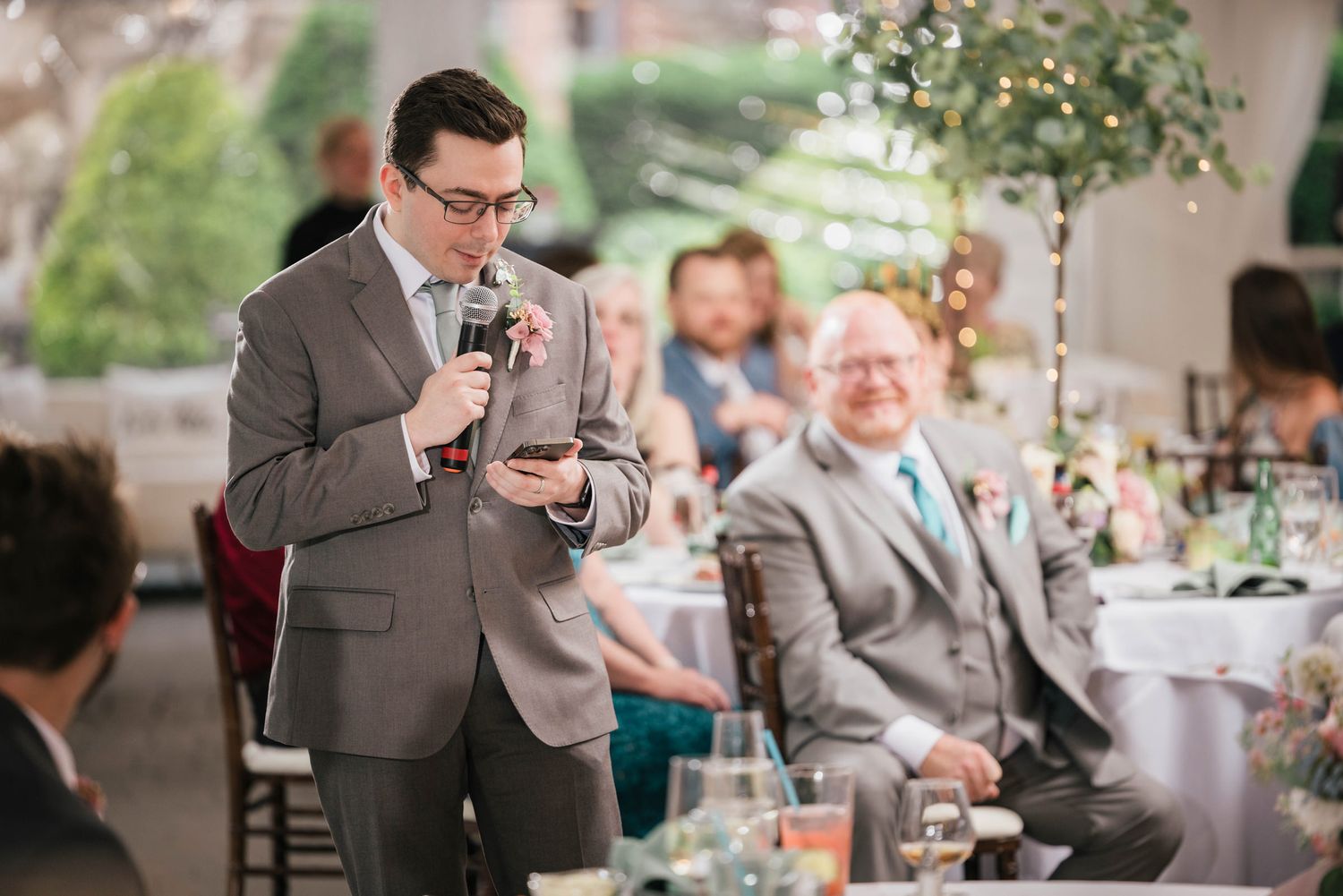 Groomsman checking his phone mid-speech, groom's father smiling behind him.