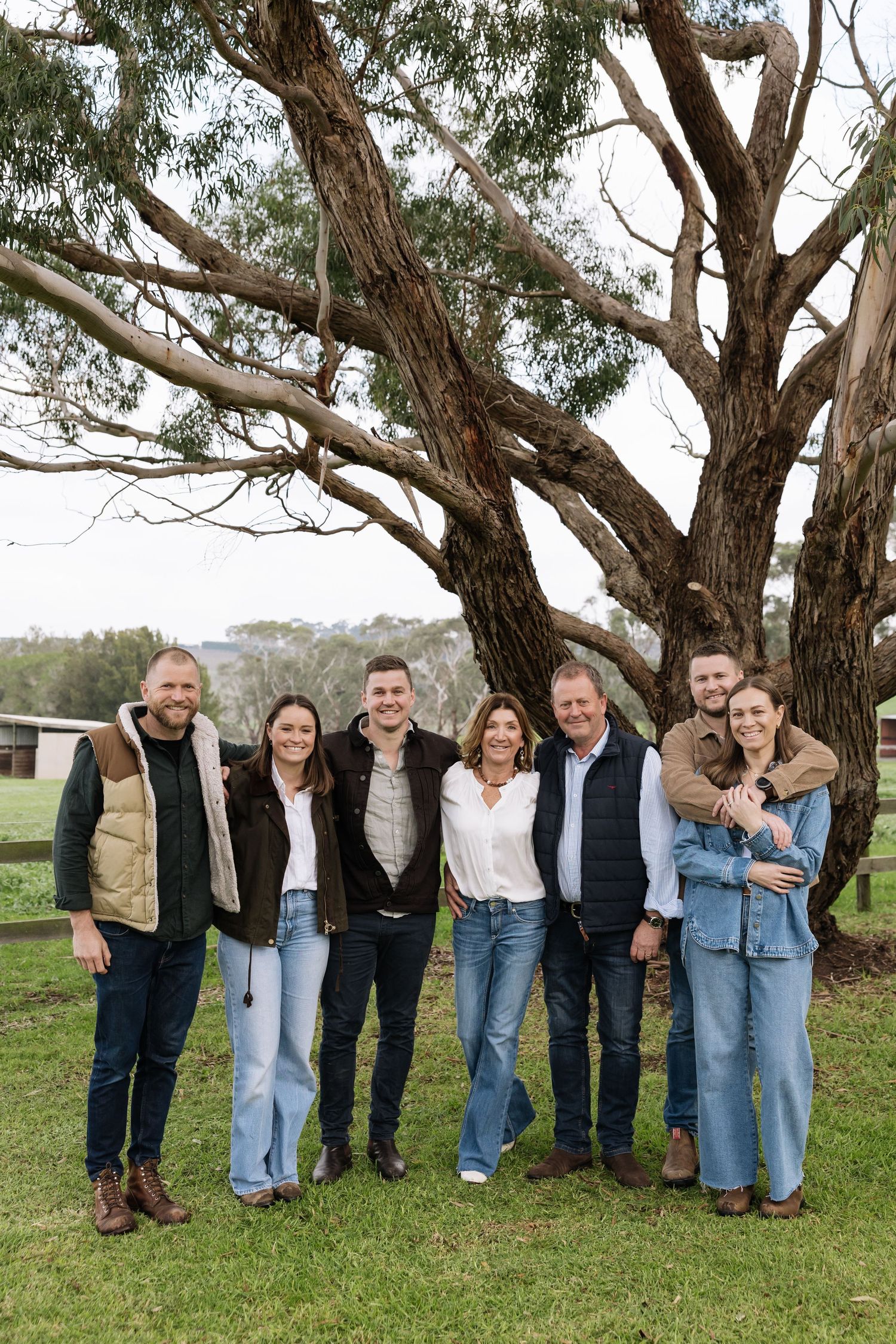 A family of seven poses together outdoors on a farm, standing in front of a large eucalyptus tree on green grass.