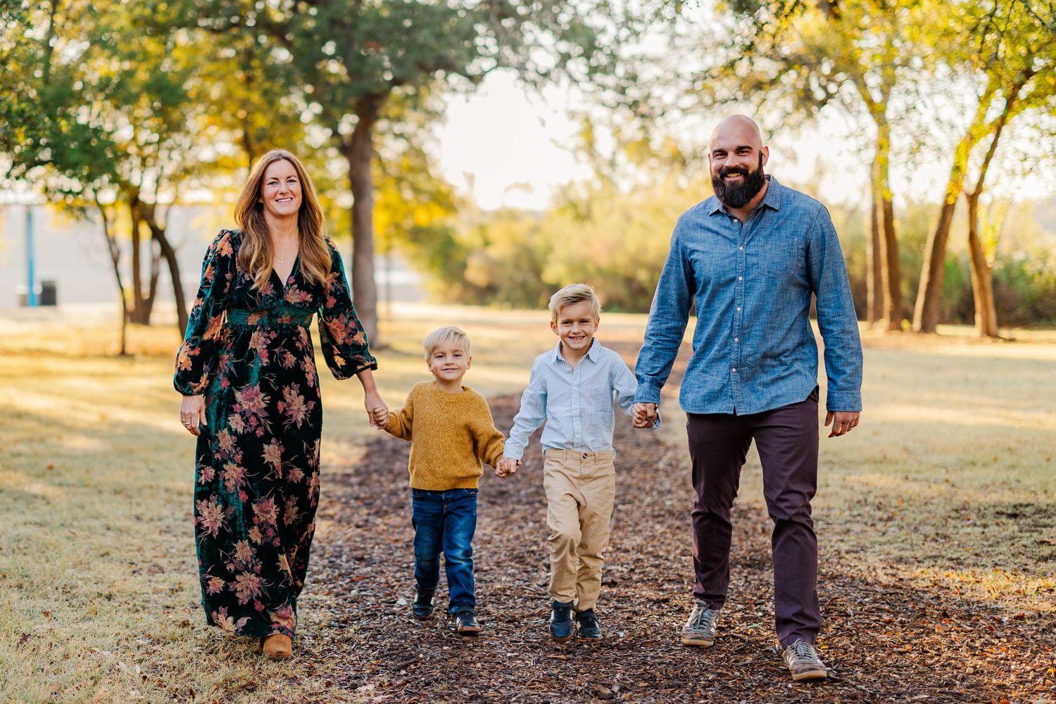 Family of four holding hands walking through a sunlit park with autumn trees in the background.