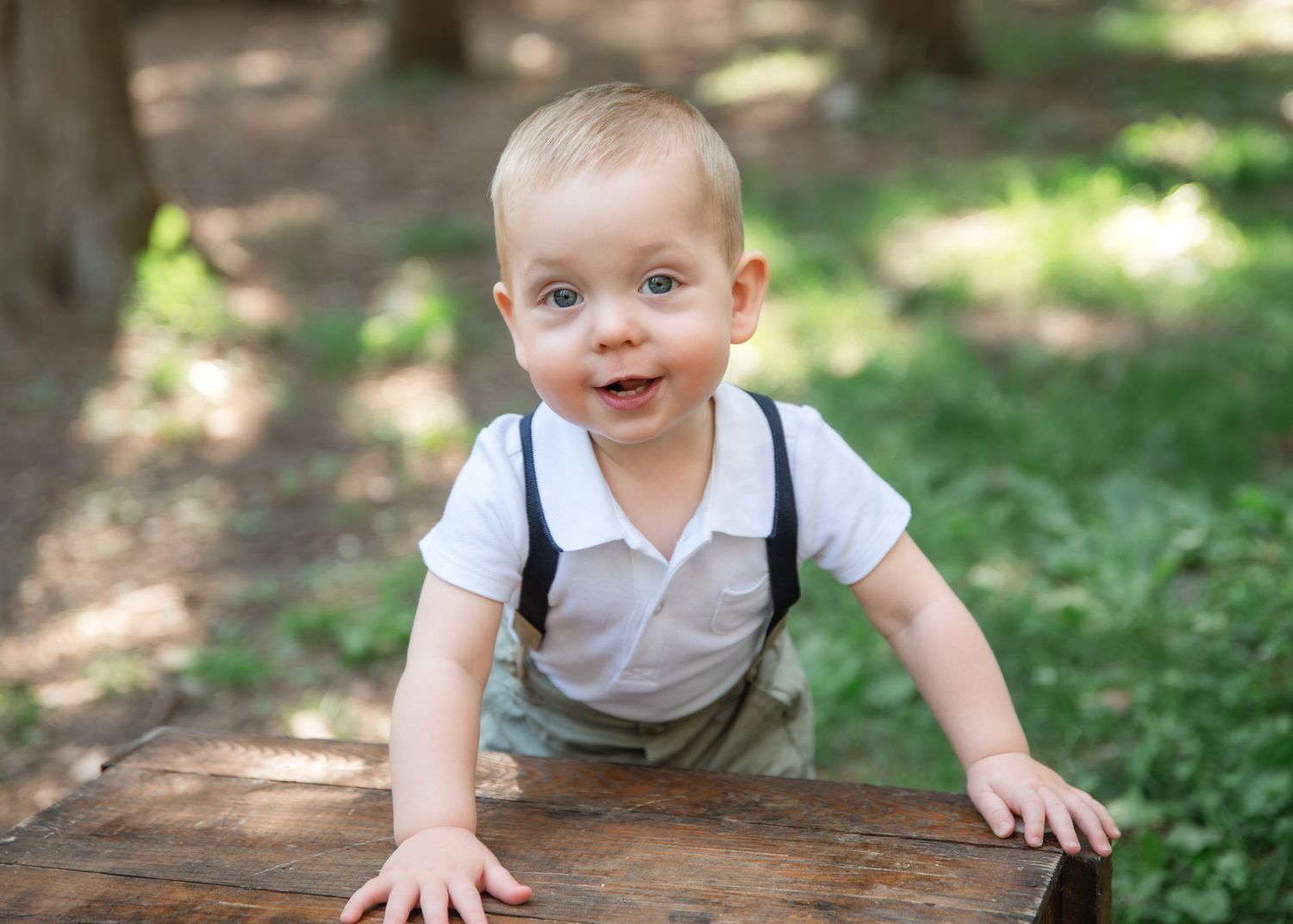 Smiling baby boy in white shirt and suspenders crawling on wooden surface outdoors in green park setting.
