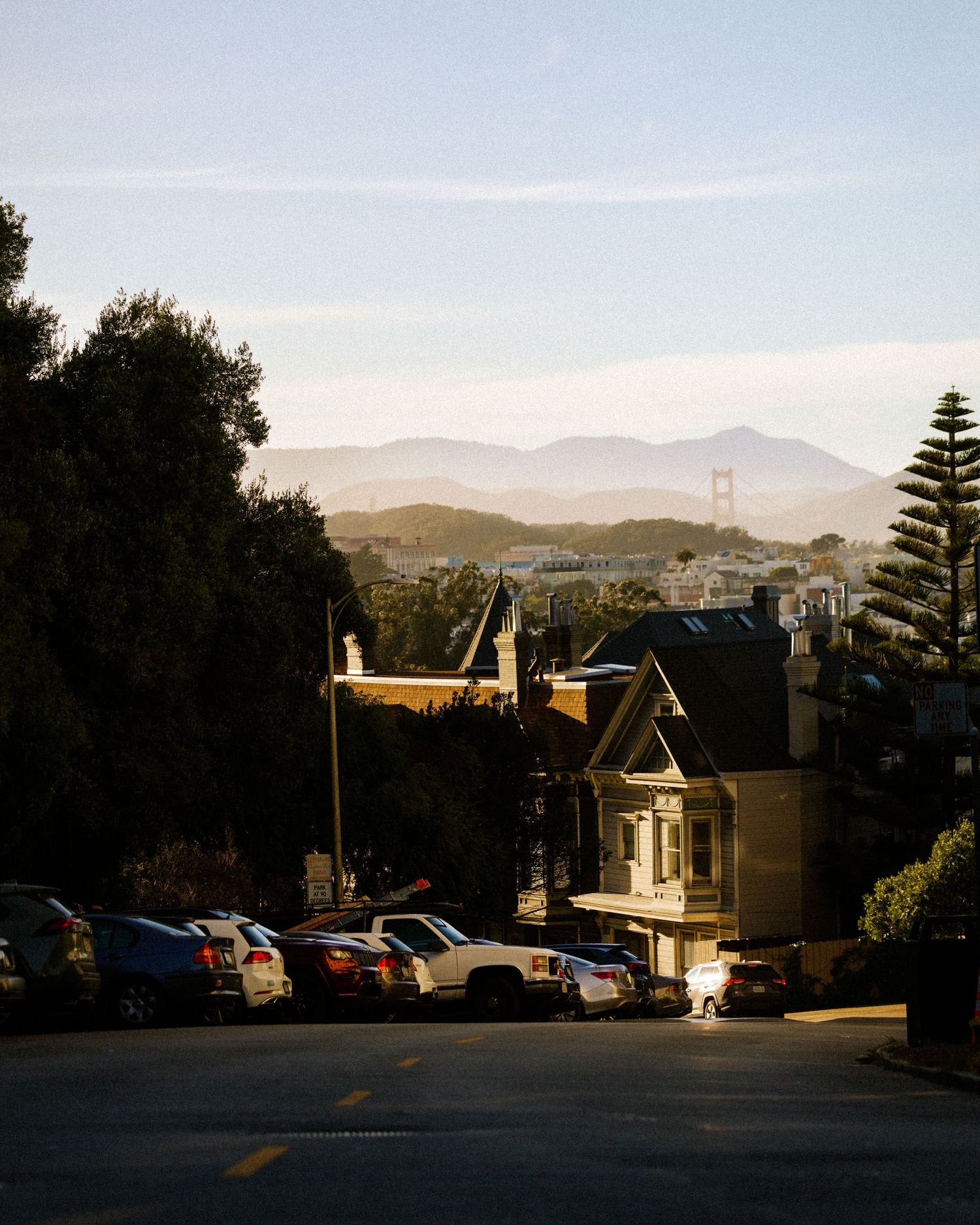 San Francisco street view with Victorian house, parked cars, Golden Gate Bridge, and mountains in background.