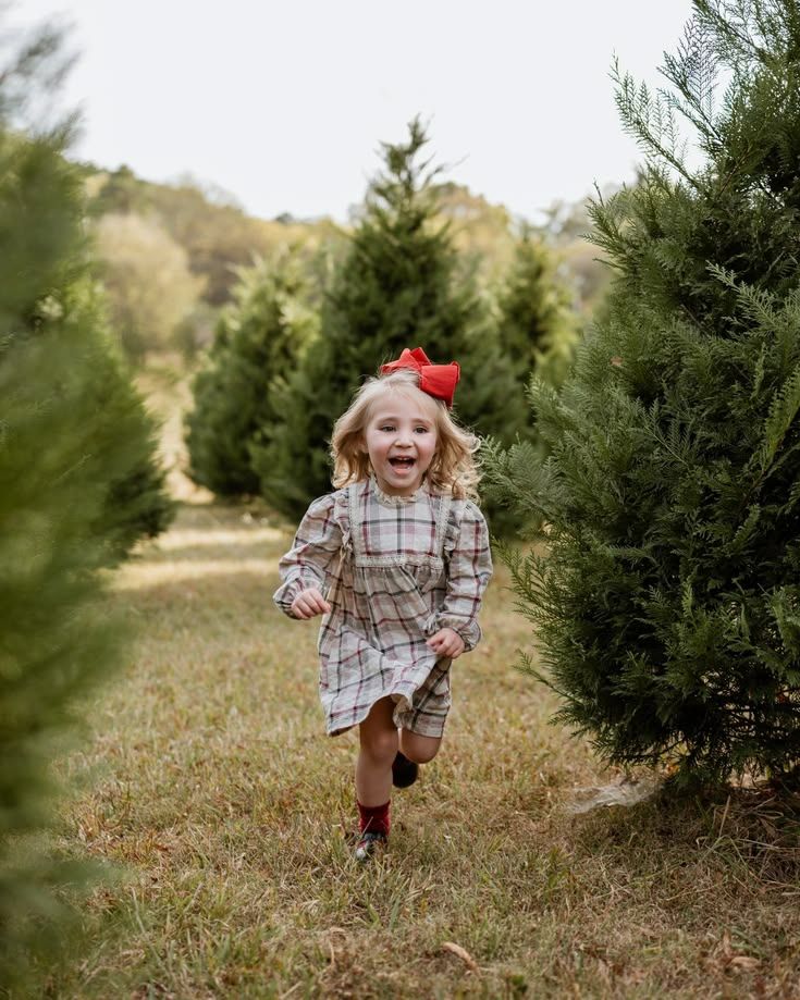 A joyful young girl in a plaid dress and red bow runs through a lush Christmas tree farm.