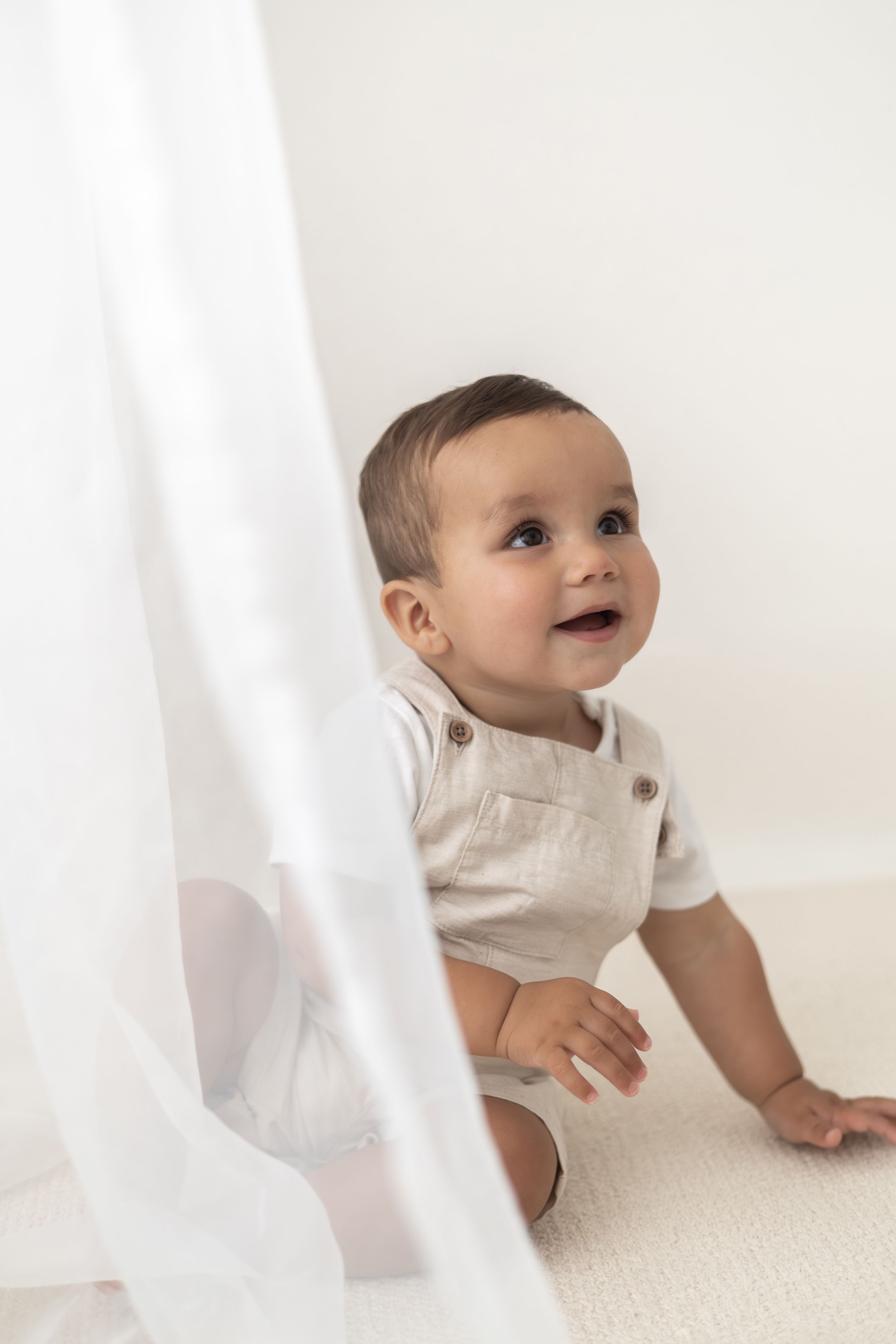 A curious baby in a linen romper crawls on a white carpet, gazing upward with a joyful smile simple neutral first birthday shoot Nottingham