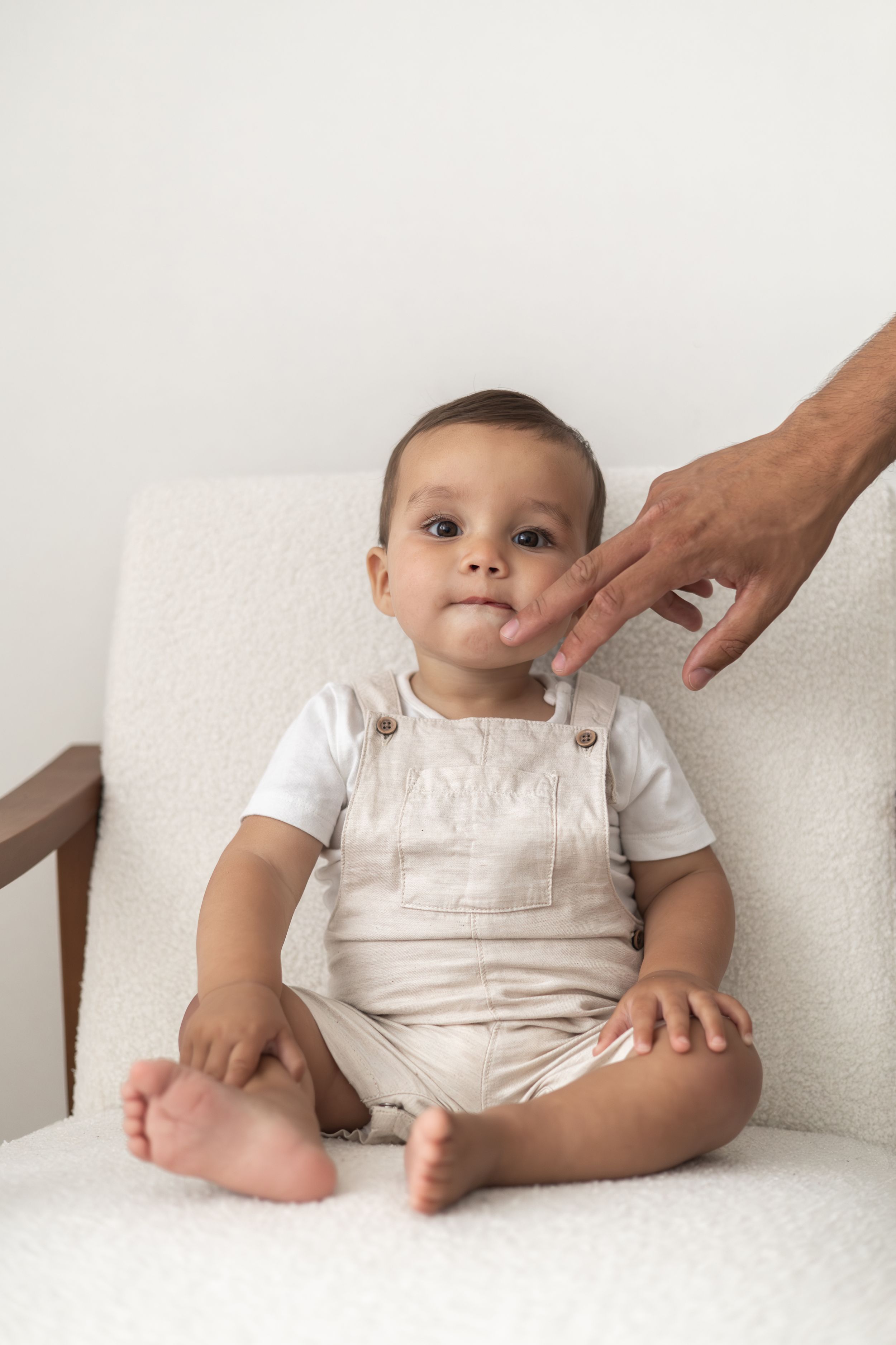 A cute baby in beige dungarees sits on a white chair as dad gently touches their chin simple neutral first birthday shoot Nottingham