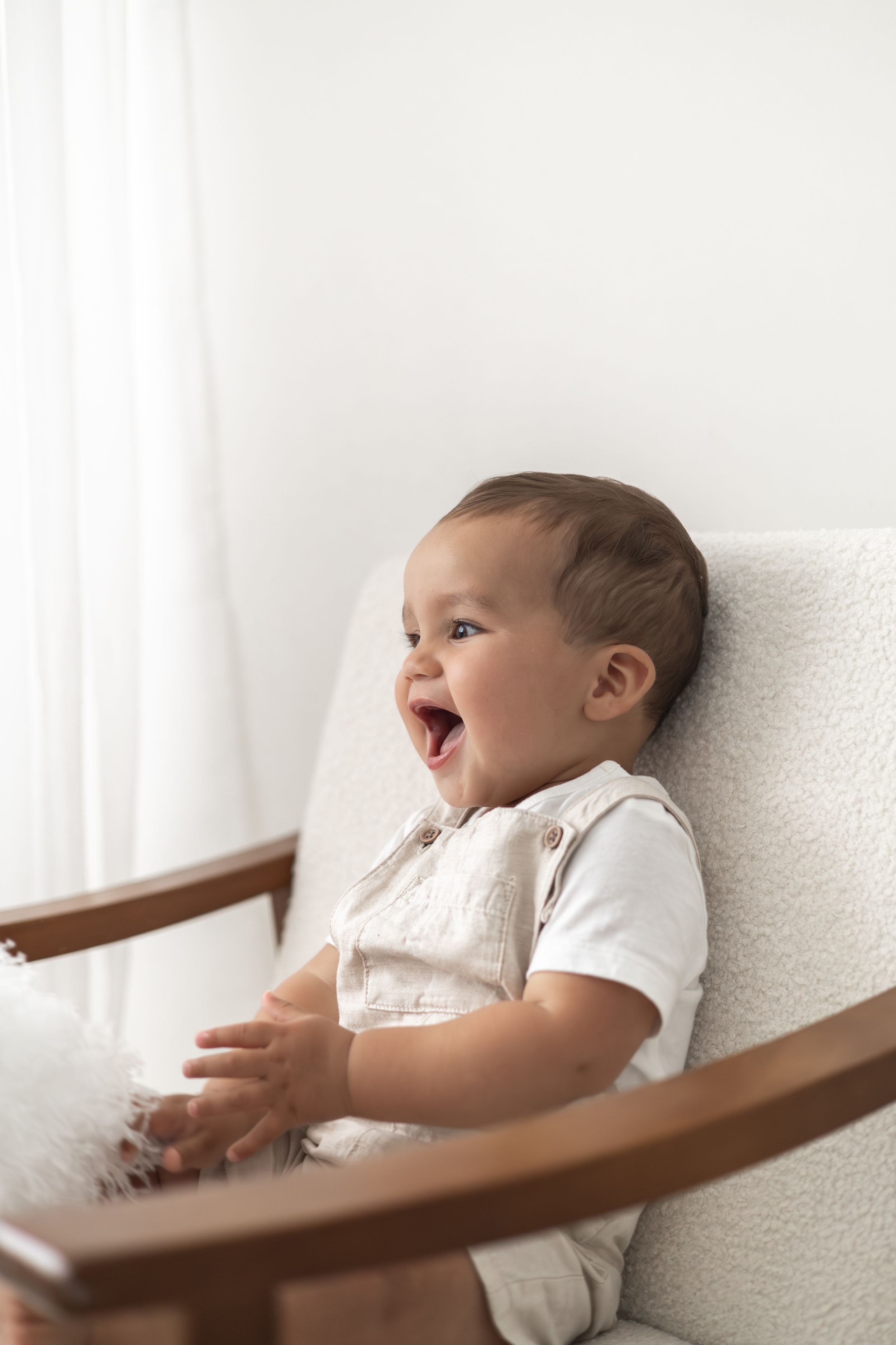 A joyful baby in beige dungarees laughs while sitting in a white wooden chair near a window simple neutral first birthday shoot Nottingham