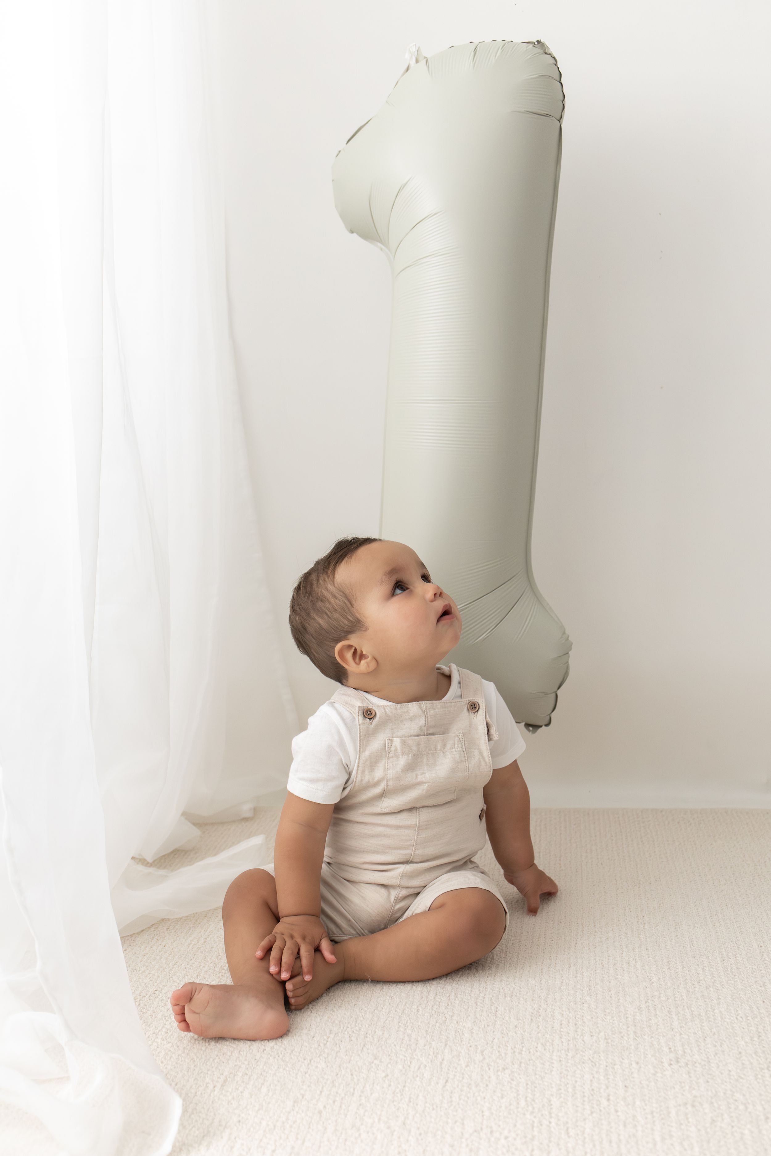 A baby in a beige romper sits on white carpet looking up at a number 1 balloon for their first birthday simple neutral first birthday Notts