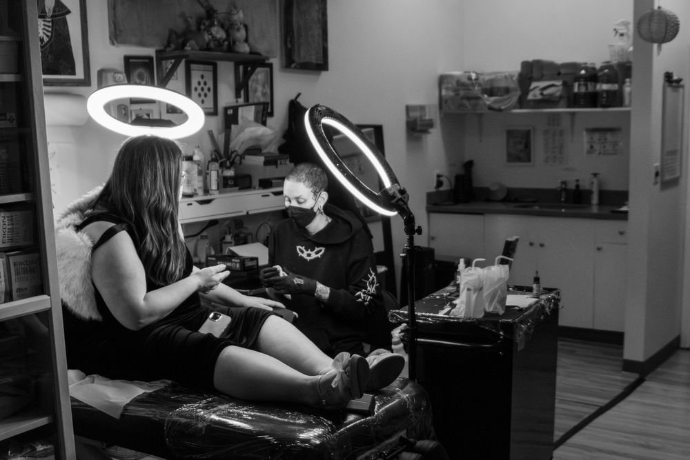 Tattoo artist works on a wedding ring tattoo, wide view of the shop, shelves and art. client and artist are framed in light halos.