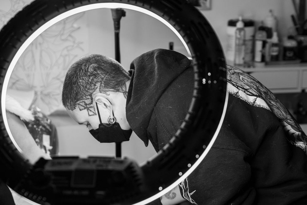 Black and white candid image of a tattooist working on a wedding ring tattoo, side view through a light ring.