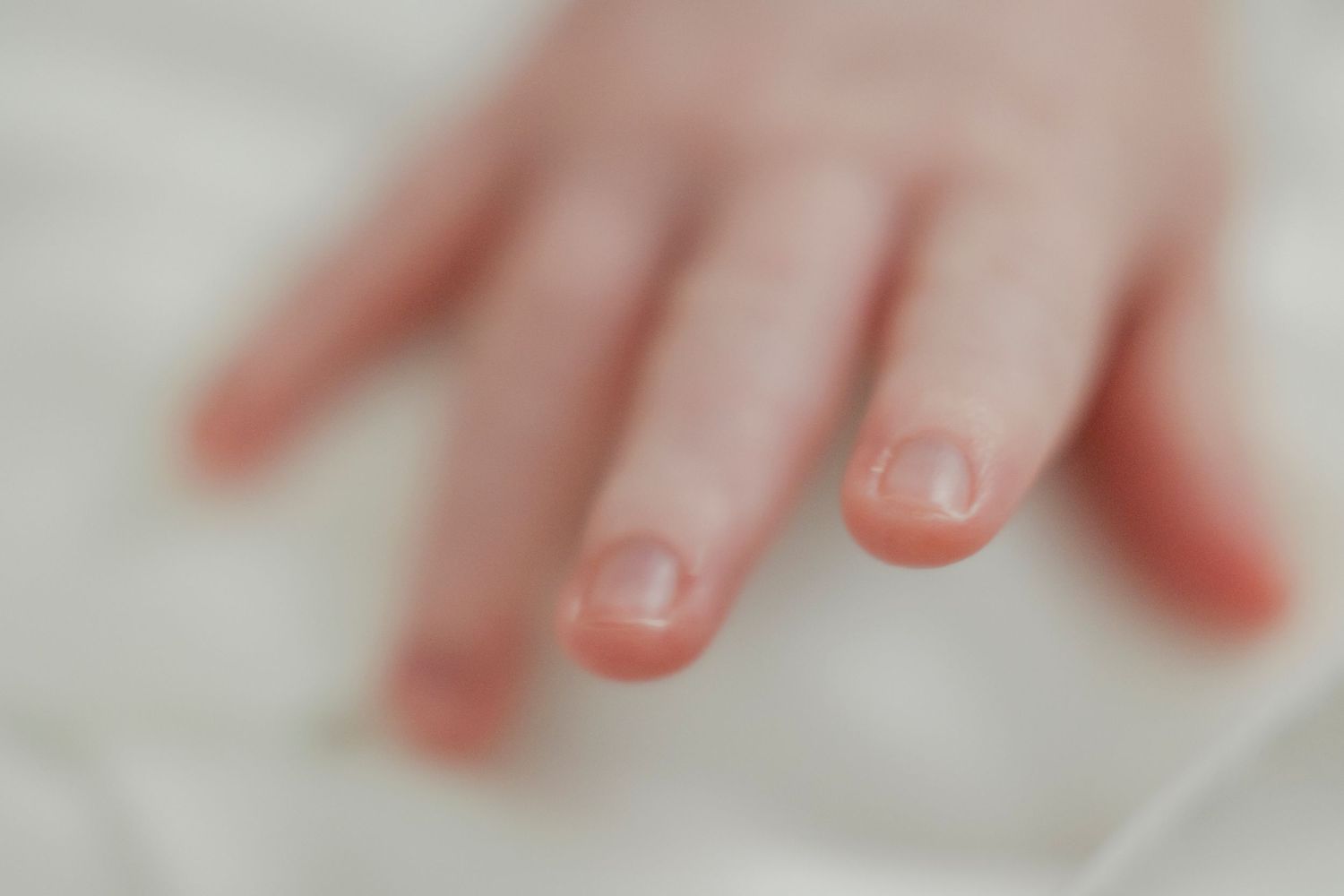 Close-up of a baby's tiny hand with delicate fingers and small nails resting on a soft white surface.