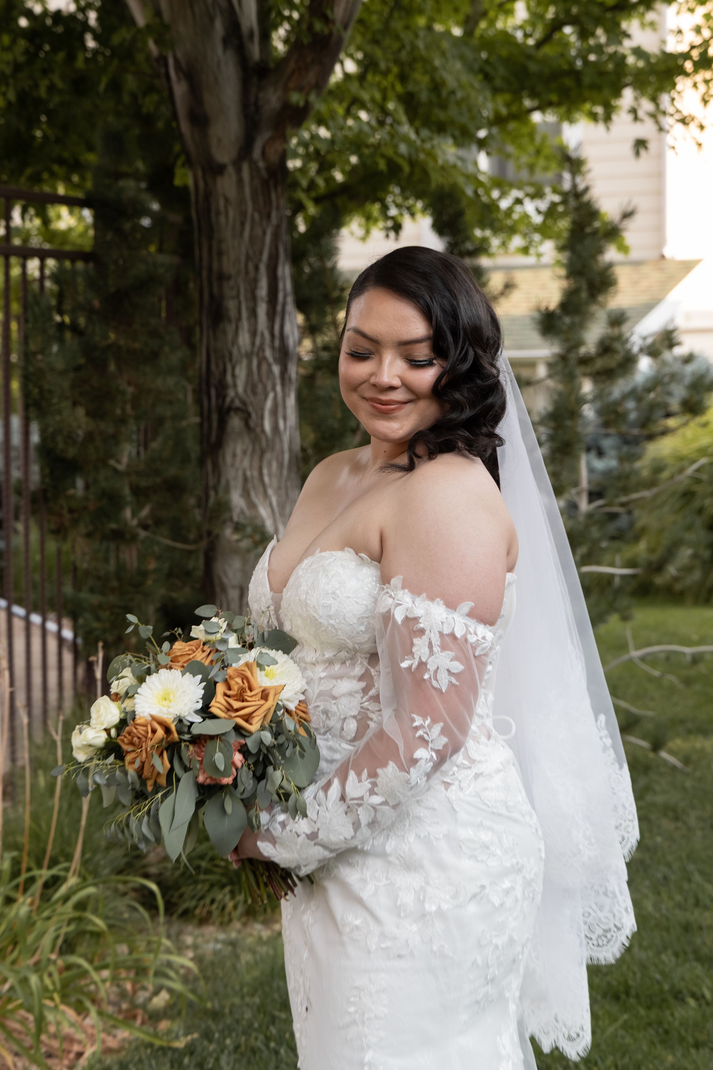 Smiling bride in off-shoulder lace gown holds orange and white bouquet outdoors under large tree.
