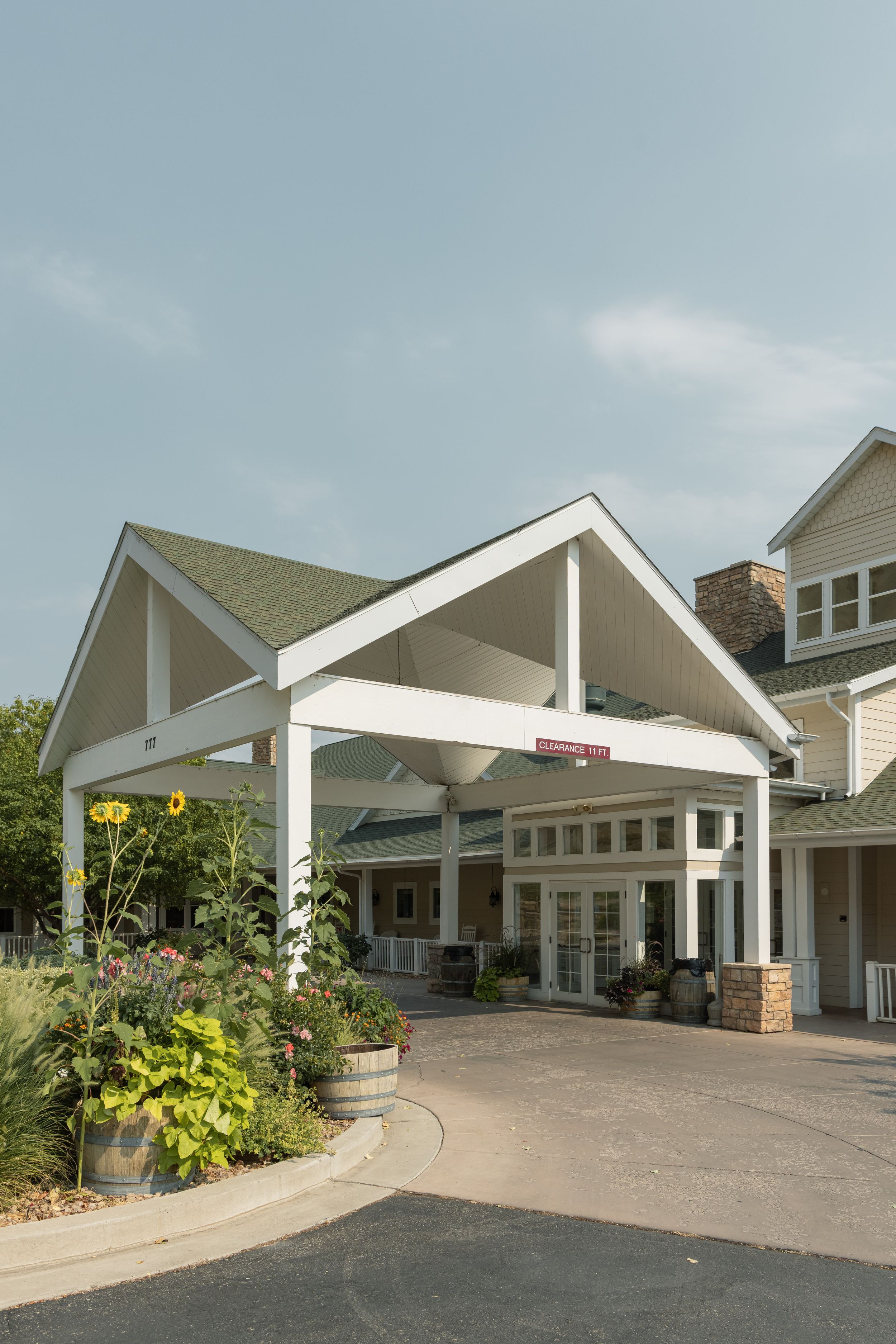 Welcoming hotel entrance with white covered portico, lush sunflowers, and manicured landscaping under a clear blue sky.