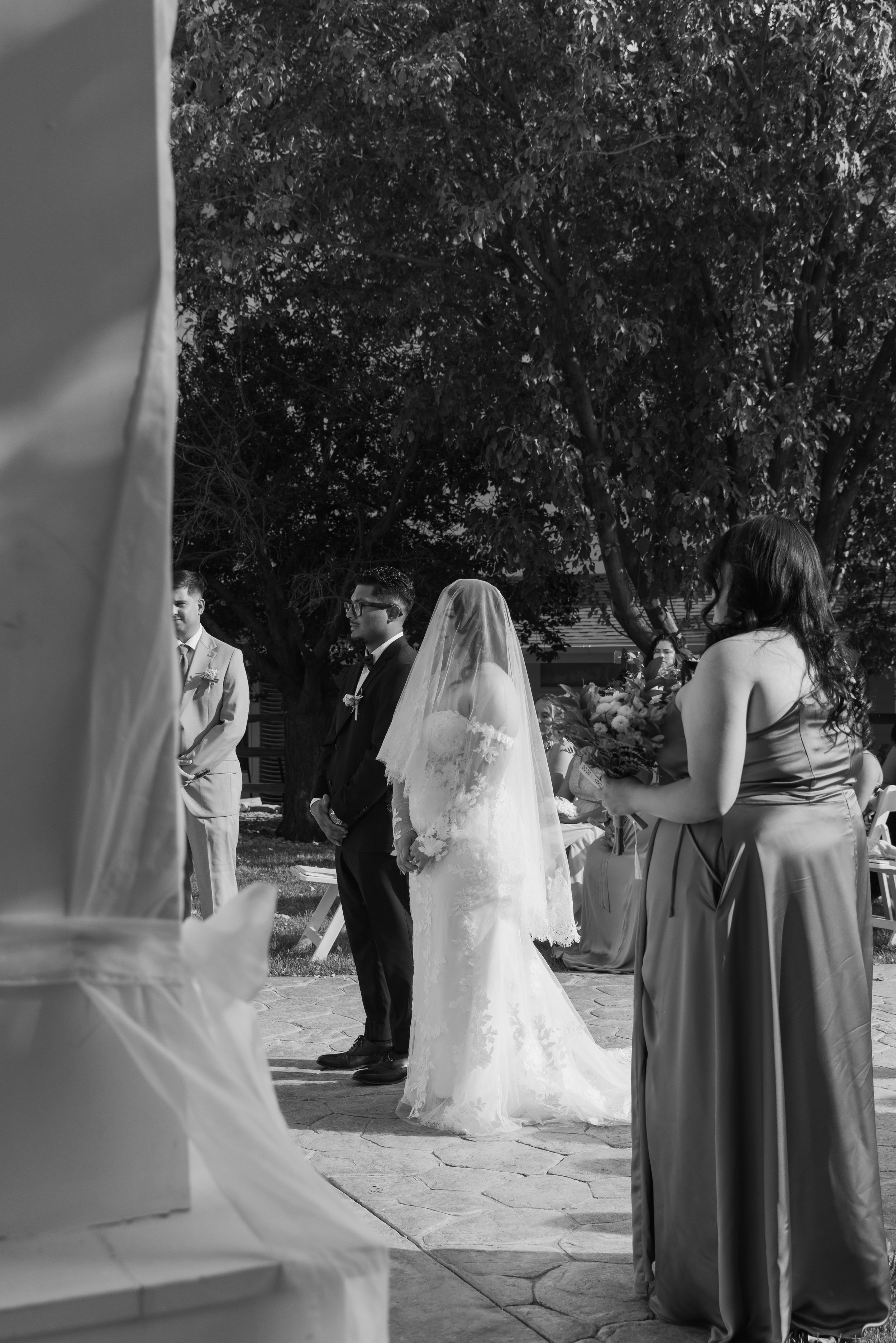 Black and white outdoor wedding ceremony with bride in veil, groom in suit, and bridesmaid holding bouquet.