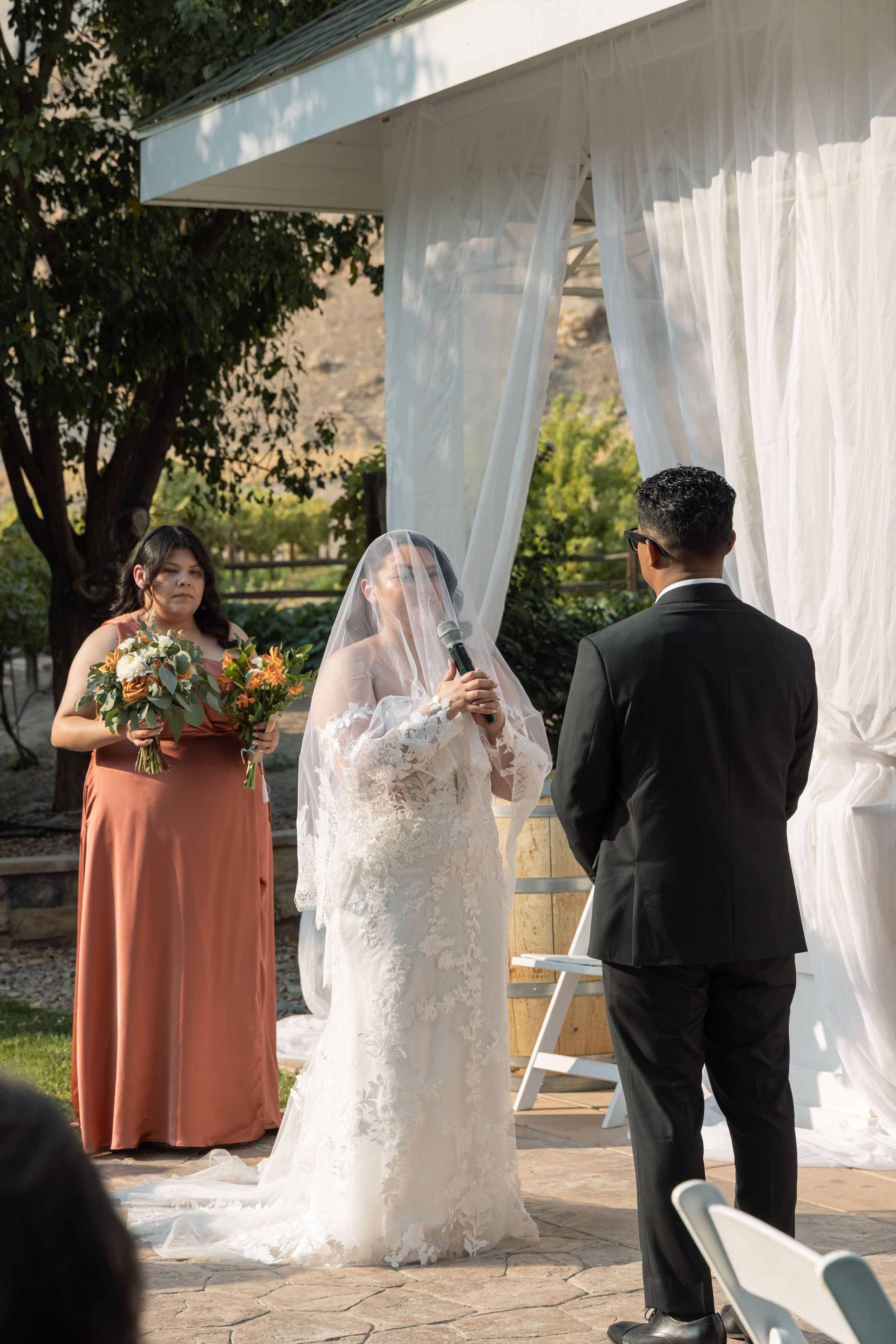 Bride in lace gown and veil exchanges vows with groom outdoors, bridesmaid in rust dress holds bouquet nearby.