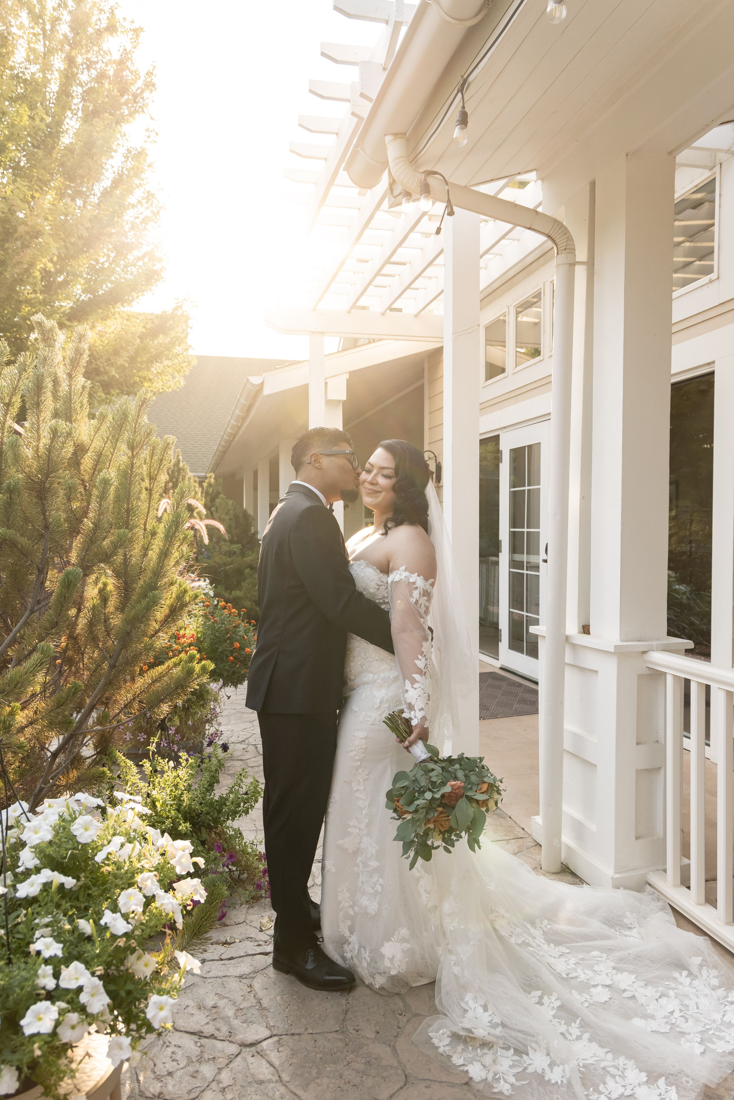 Bride in lace gown and groom in black tuxedo share a romantic moment outside an elegant venue at golden hour.