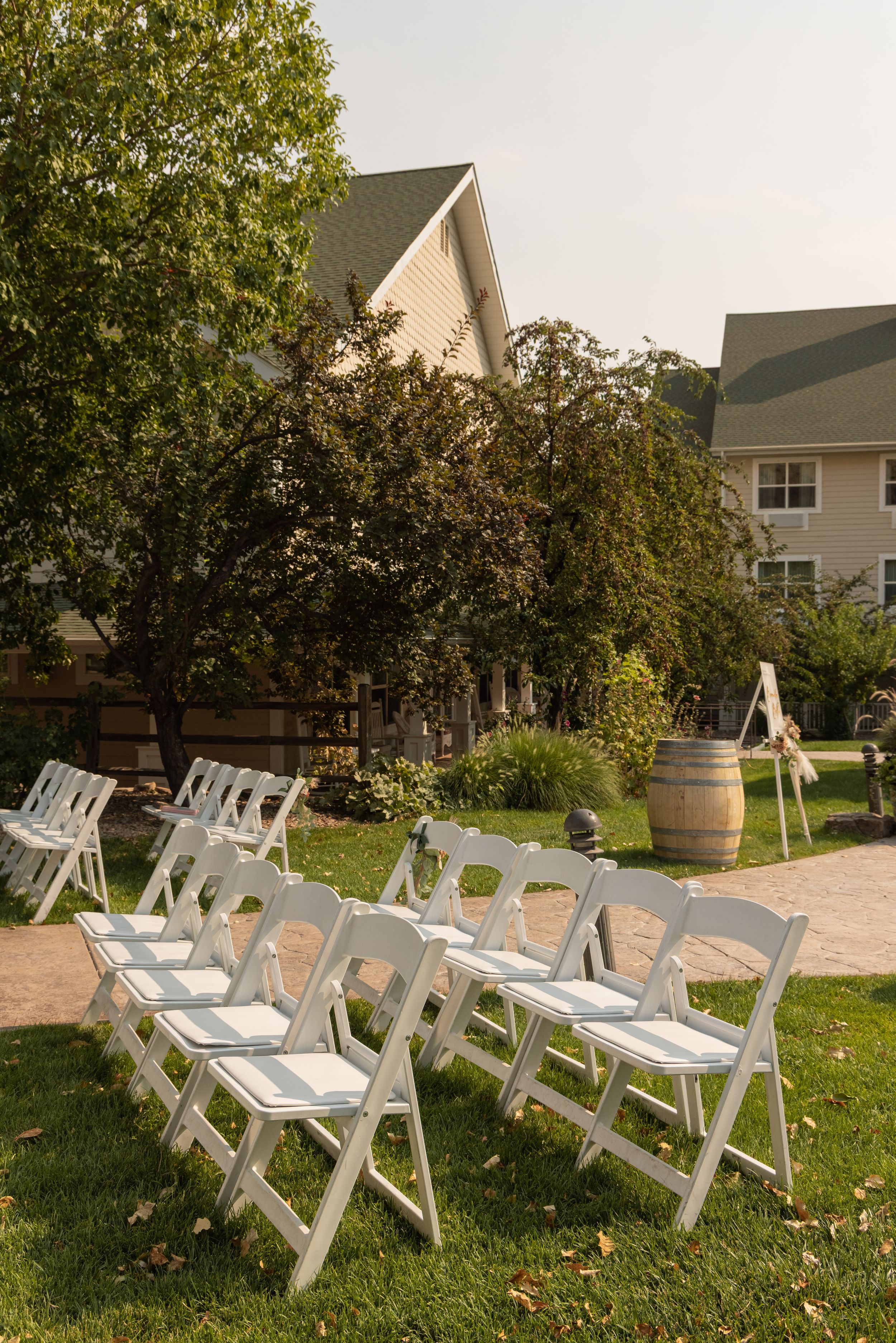 White folding chairs arranged on green lawn for an outdoor wedding ceremony near a wooden barrel and house.