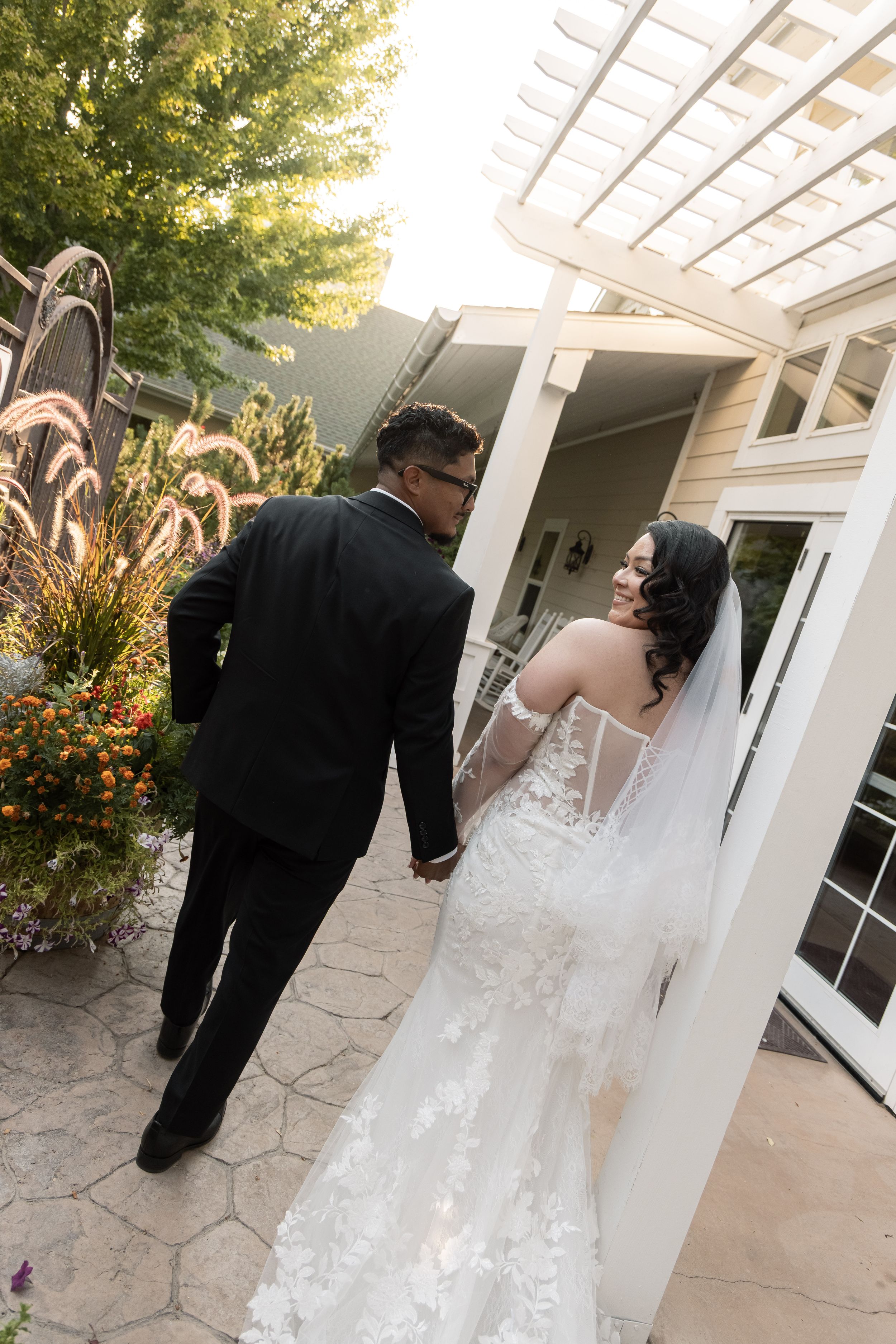 Bride in lace gown and veil holds hands with groom in black tuxedo walking through elegant outdoor wedding venue.