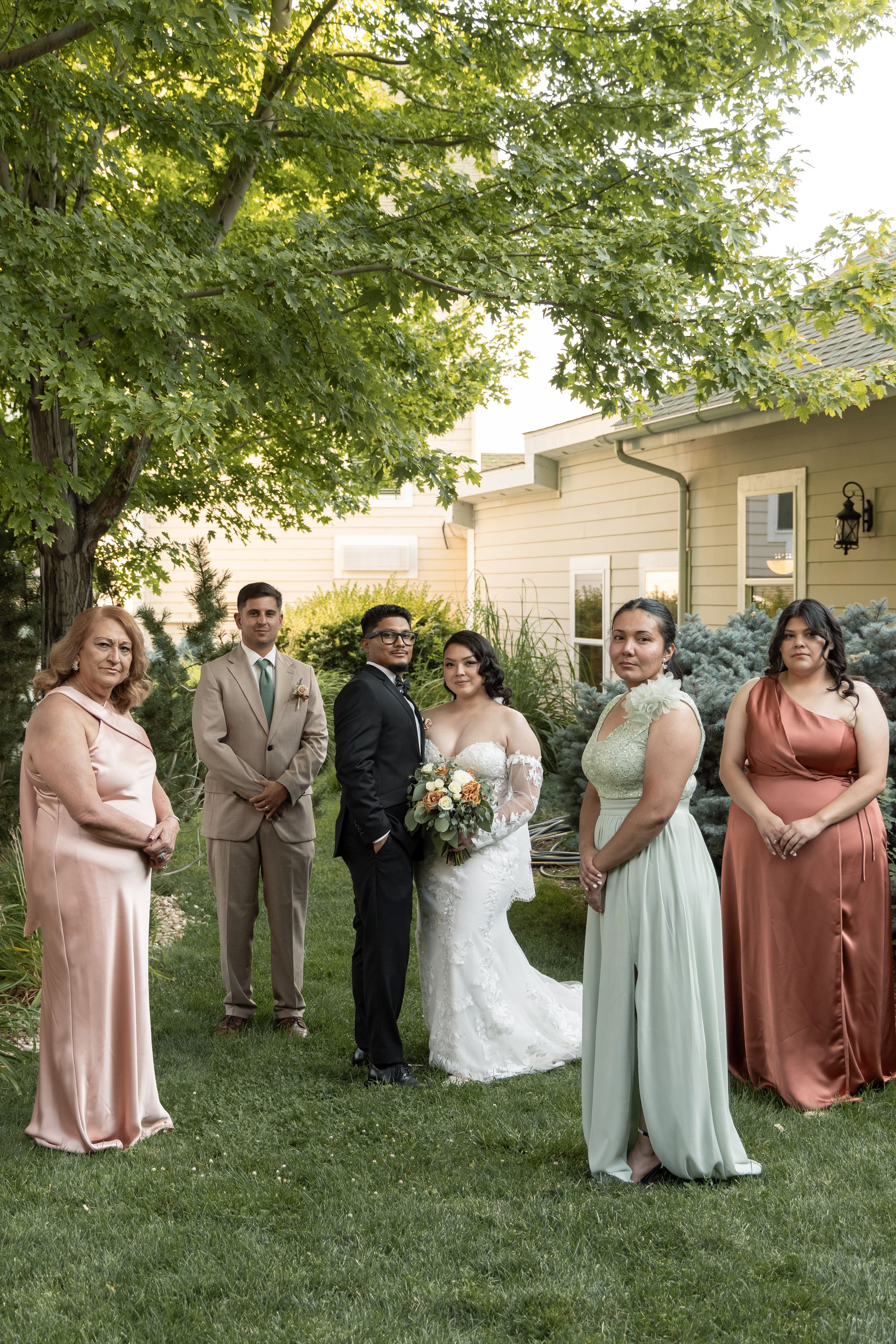Wedding party poses outdoors on green lawn, bride in lace gown with bouquet, groom in black suit, attendants in sage and dusty rose gowns.
