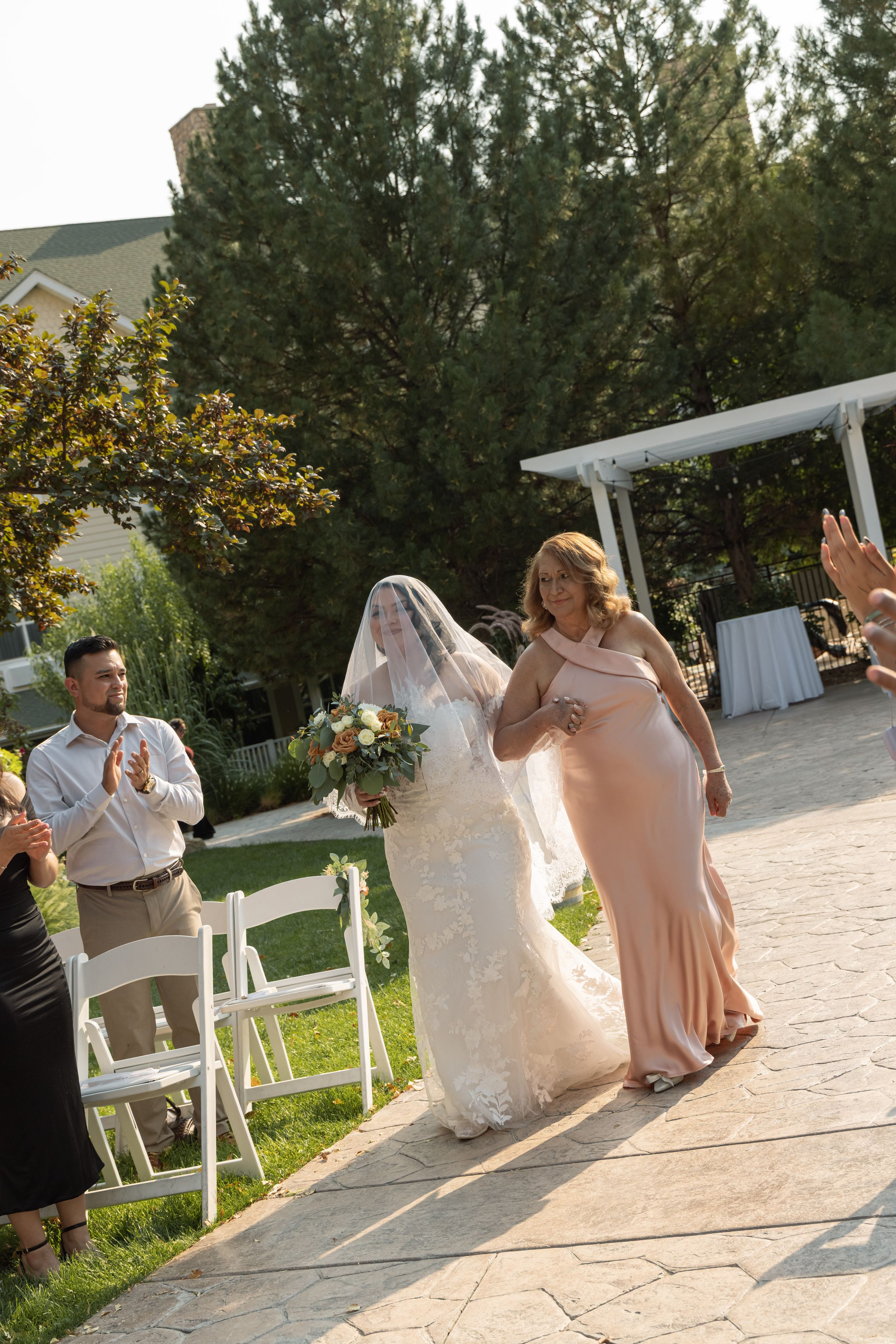 Bride in white lace gown and veil walks down outdoor aisle with woman in blush dress, guests applauding at garden wedding.