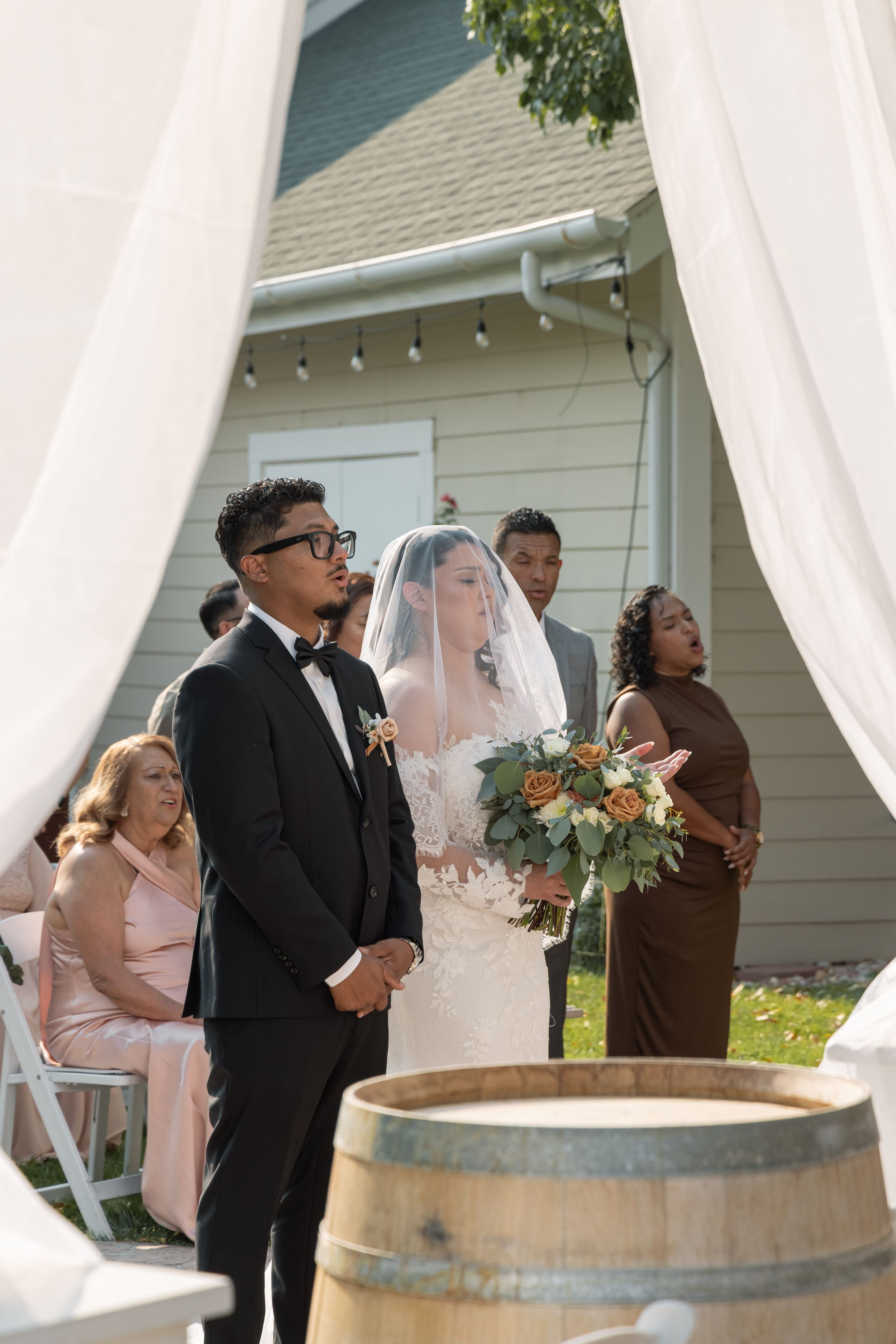 Outdoor wedding ceremony with groom in tuxedo and bride in white gown holding floral bouquet, framed by white draping near wine barrel.