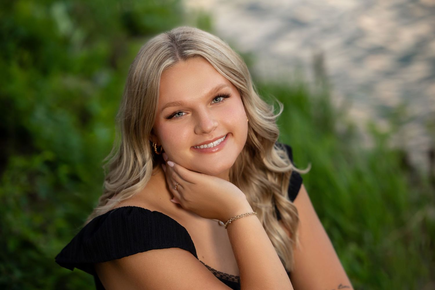 Smiling teenage girl with blonde wavy hair posing outdoors in a black off-shoulder top against green foliage backdrop.